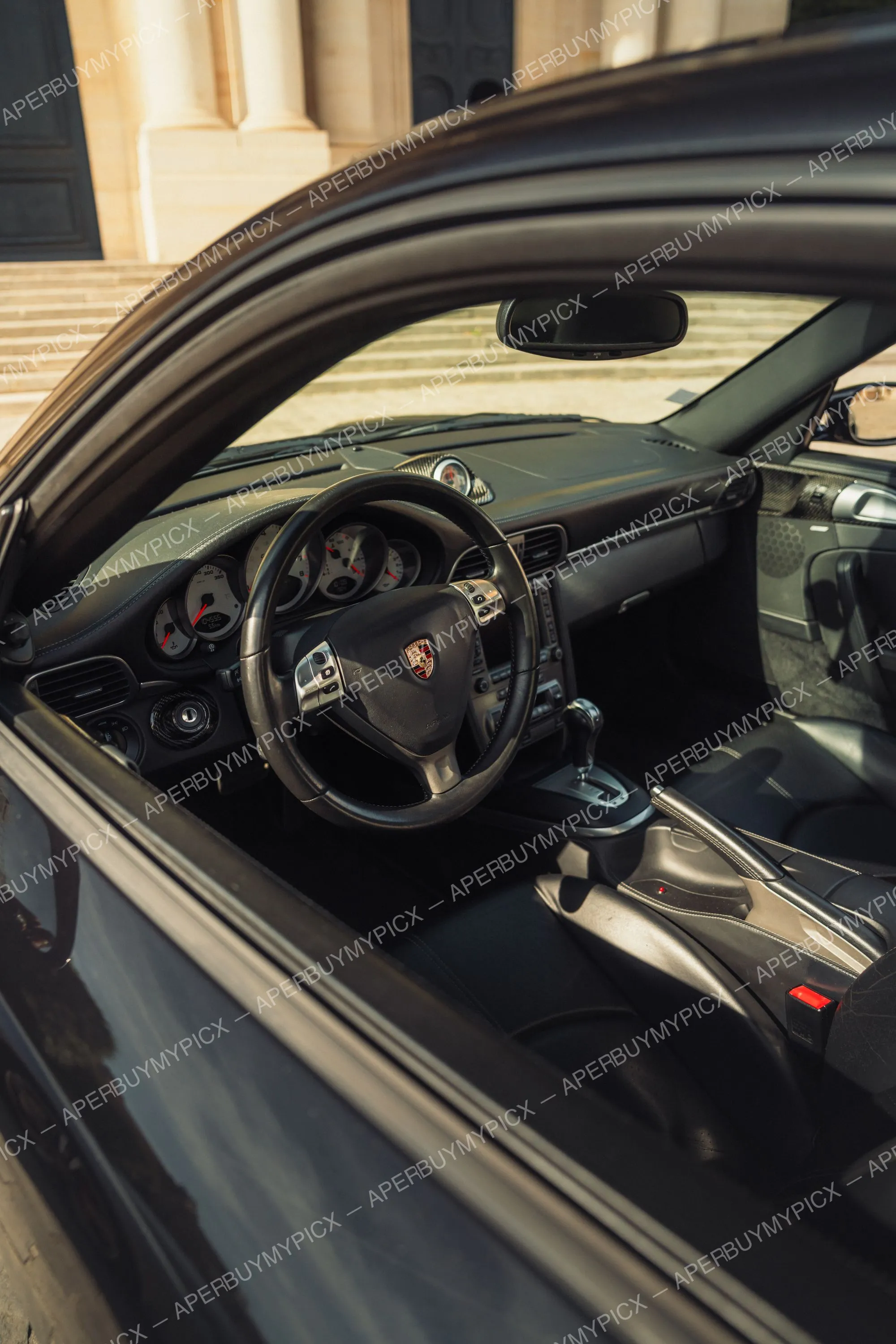Vue de l'intérieur cuir noir d'une Porsche 911 Turbo depuis la portière ouverte, avec volant à écusson Porsche, tableau de bord à compteurs analogiques et levier de vitesses Tiptronic, devant un bâtiment à colonnes.