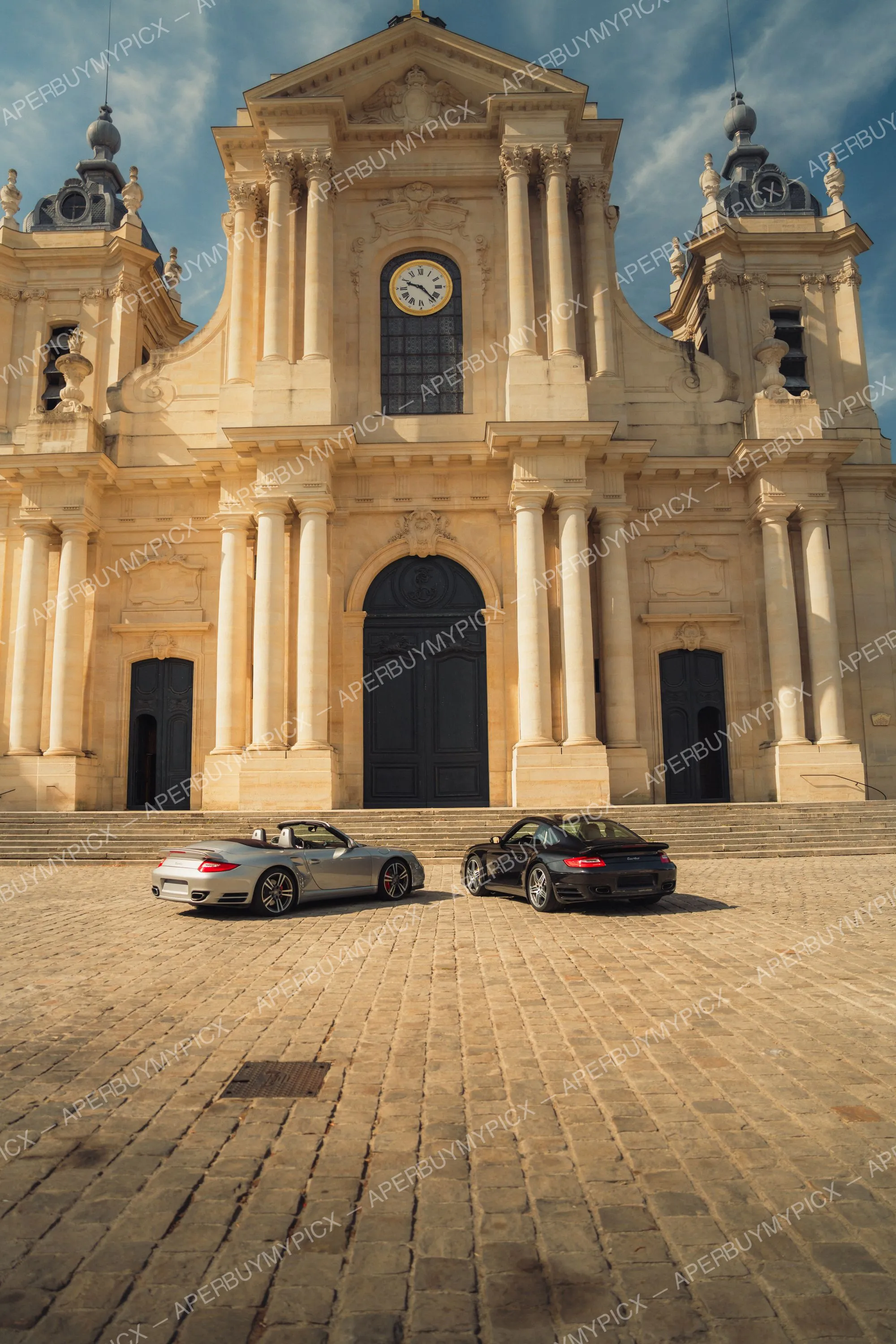 Deux Porsche 911 Turbo Cabriolet, une argentée et une noire, garées sur une place pavée devant la façade baroque d'une cathédrale avec une horloge et des colonnes.