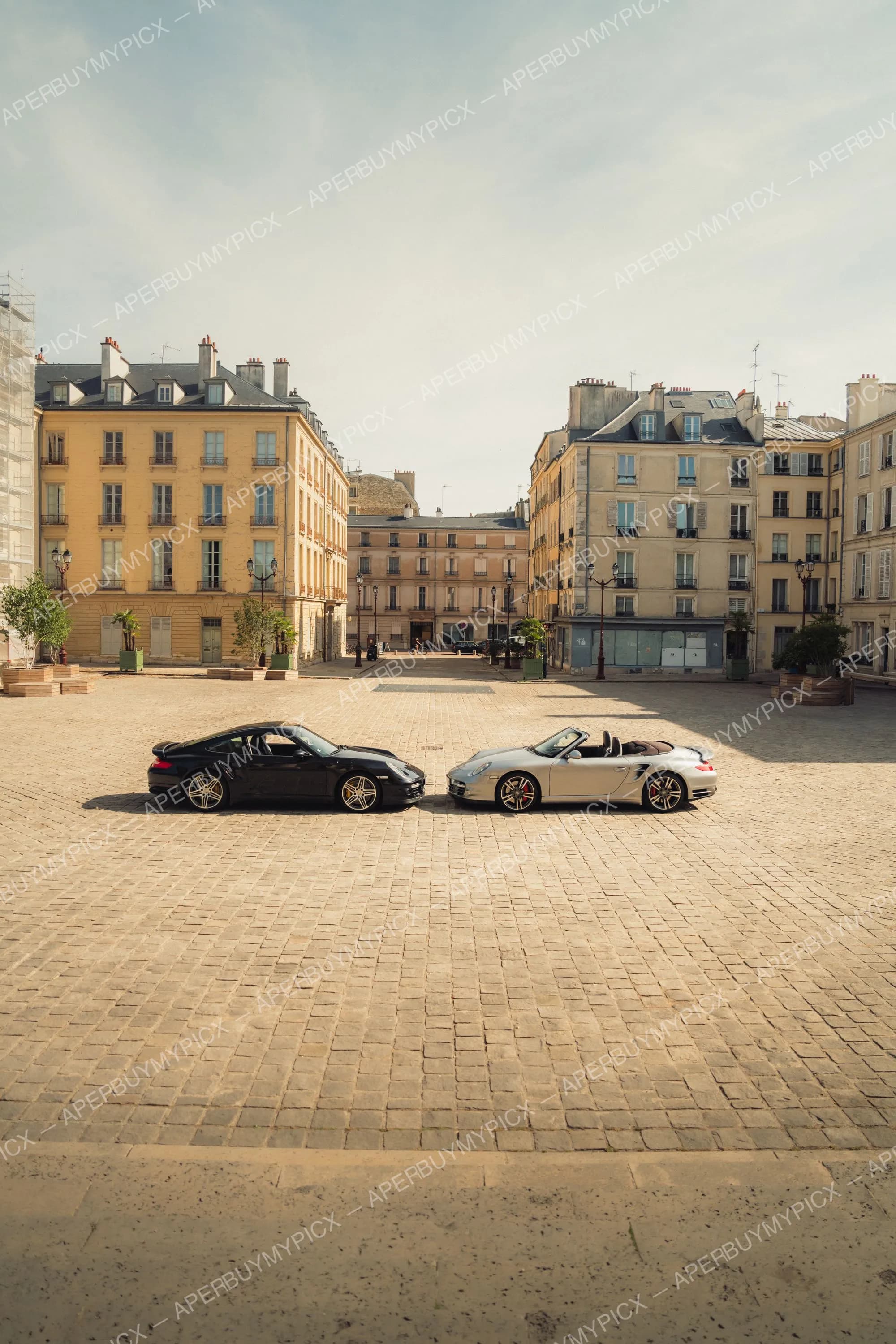 Une Porsche 911 Turbo noire coupé et une Porsche 911 Turbo grise cabriolet garées face à face sur une place pavée à Paris, entourée d'immeubles haussmanniens sous un ciel clair.