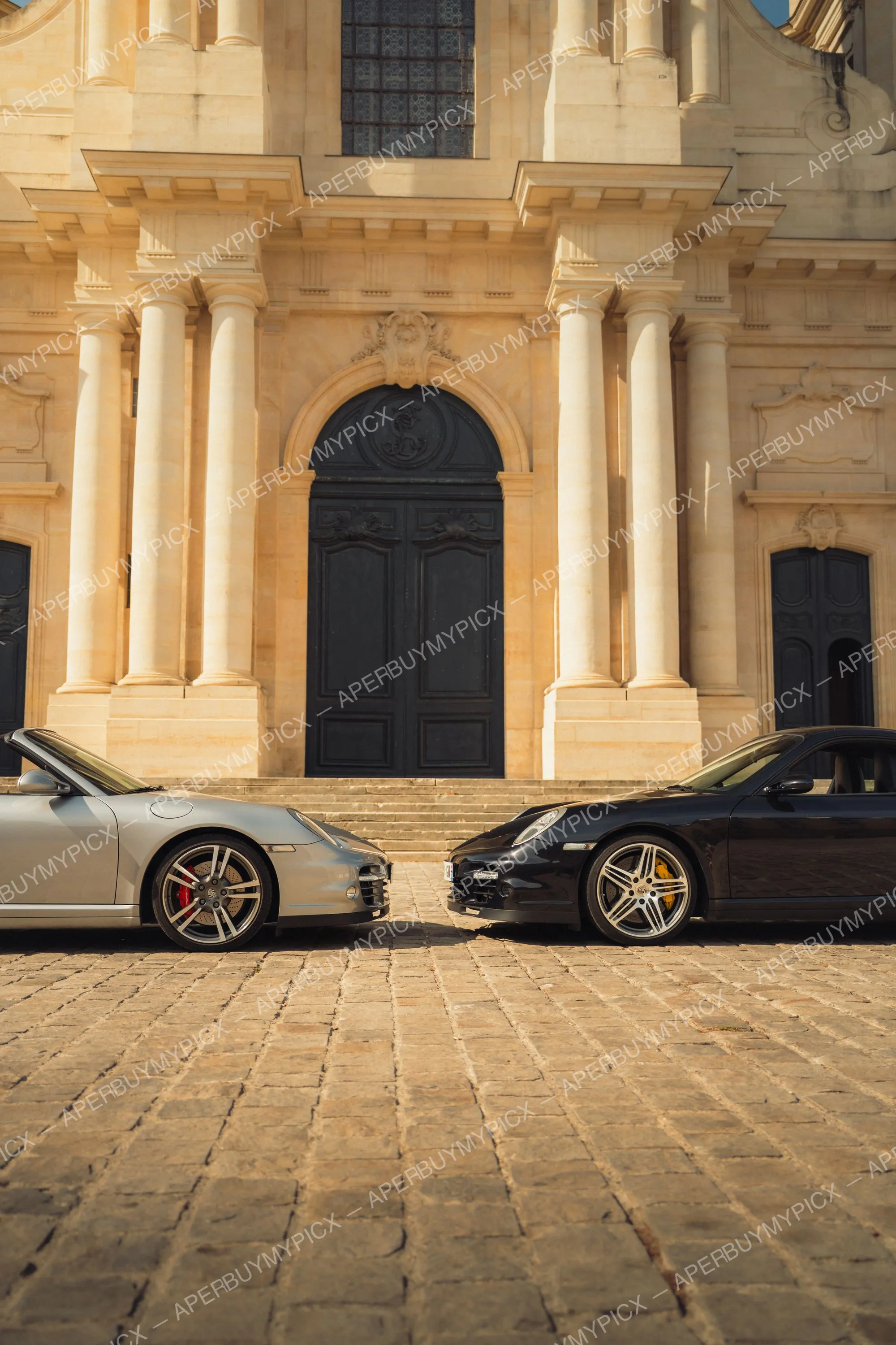 Deux Porsche 911 Turbo Cabriolet, une grise et une noire, garées face à face sur des pavés devant une imposante façade en pierre avec colonnes et grande porte noire cintrée.