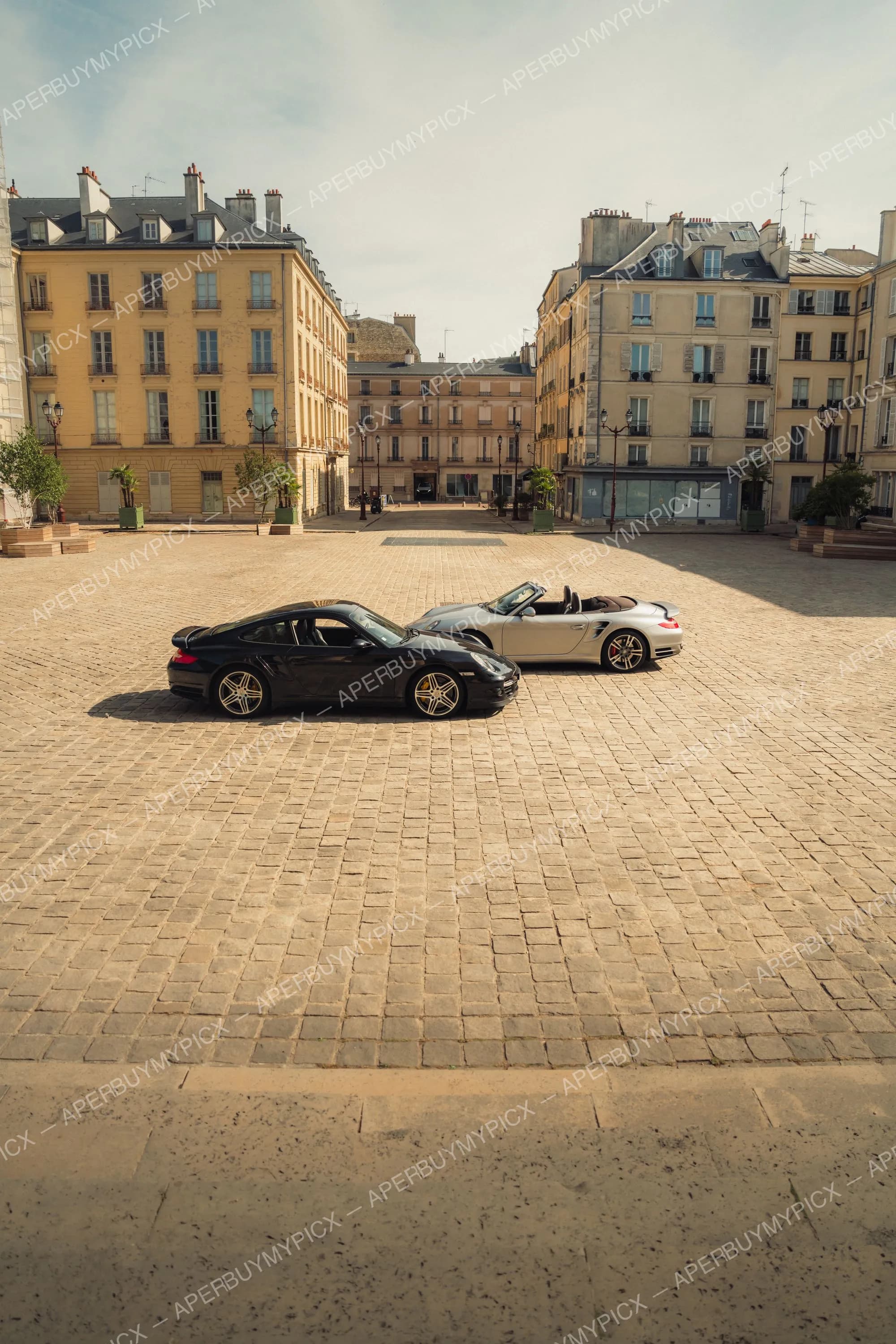 Une Porsche 911 Turbo noire coupé et une Porsche 911 Turbo cabriolet argentée garées sur une place pavée bordée d'immeubles haussmanniens à Paris sous un ciel clair.