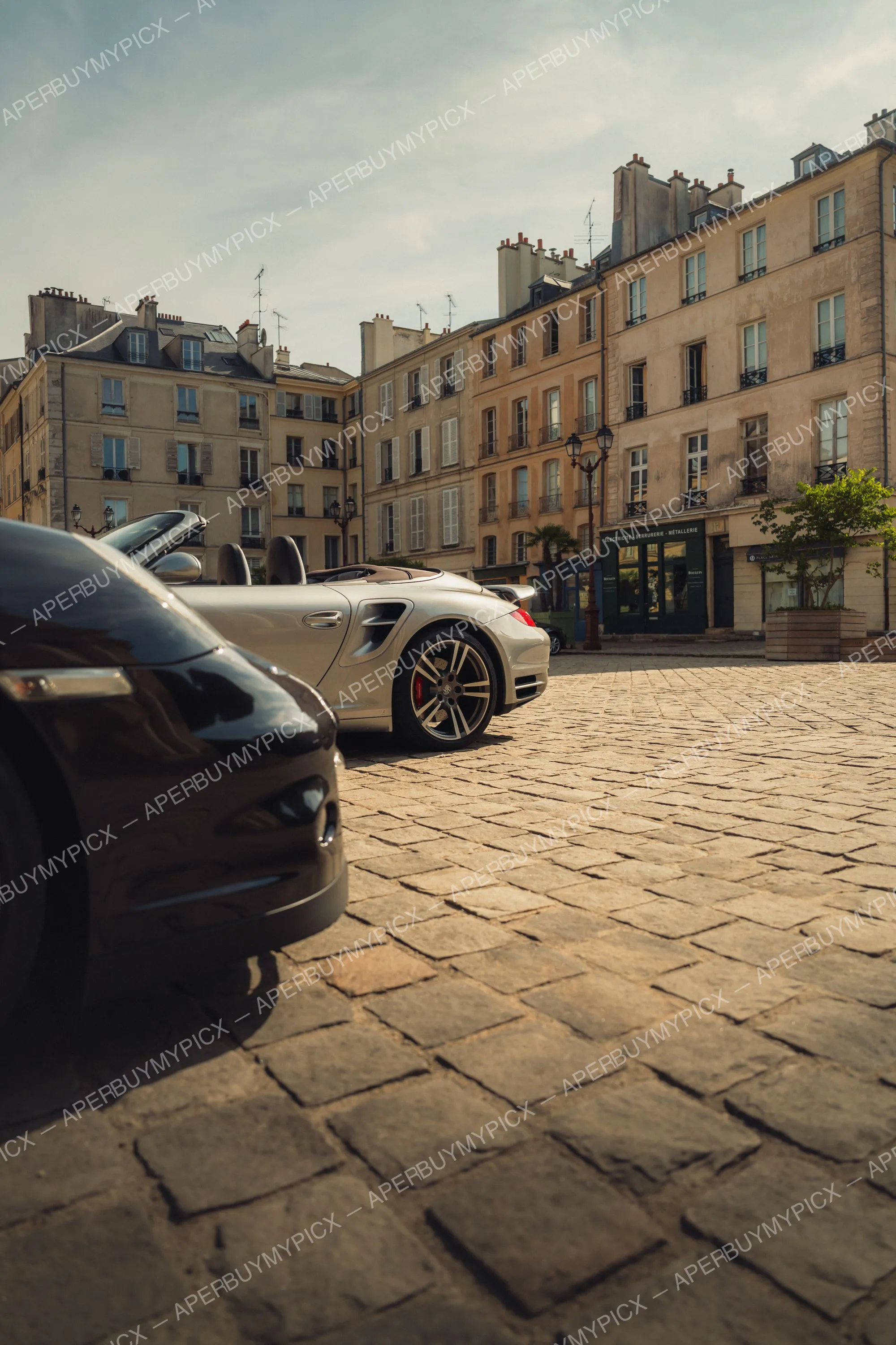 Porsche 911 Turbo cabriolet argentée garée sur une place pavée parisienne, avec une autre Porsche noire au premier plan et des immeubles haussmanniens en arrière-plan sous un ciel nuageux
