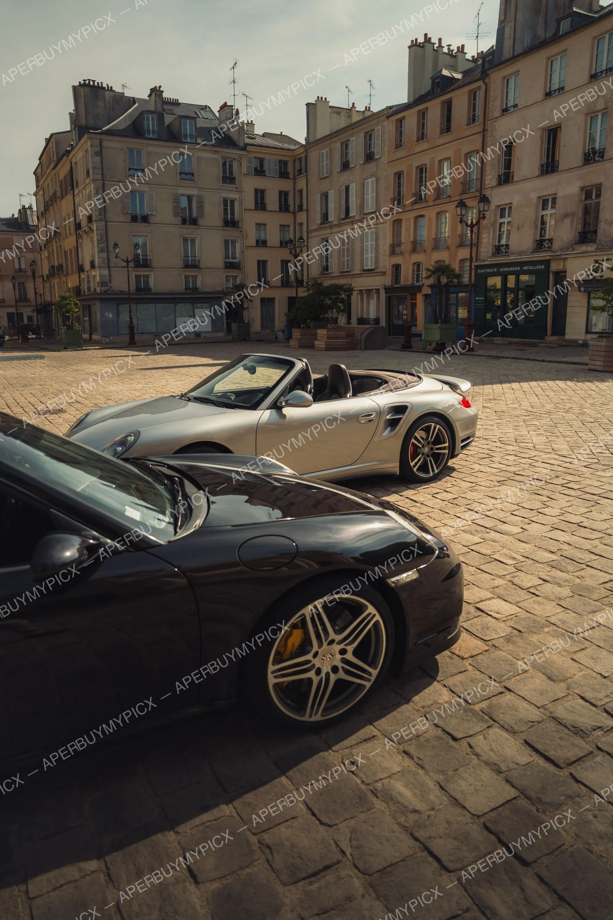 Porsche 911 Turbo Cabriolet argentée décapotée garée sur une place pavée parisienne, avec une Porsche 911 noire au premier plan et des immeubles haussmanniens en arrière-plan sous une lumière dorée.
