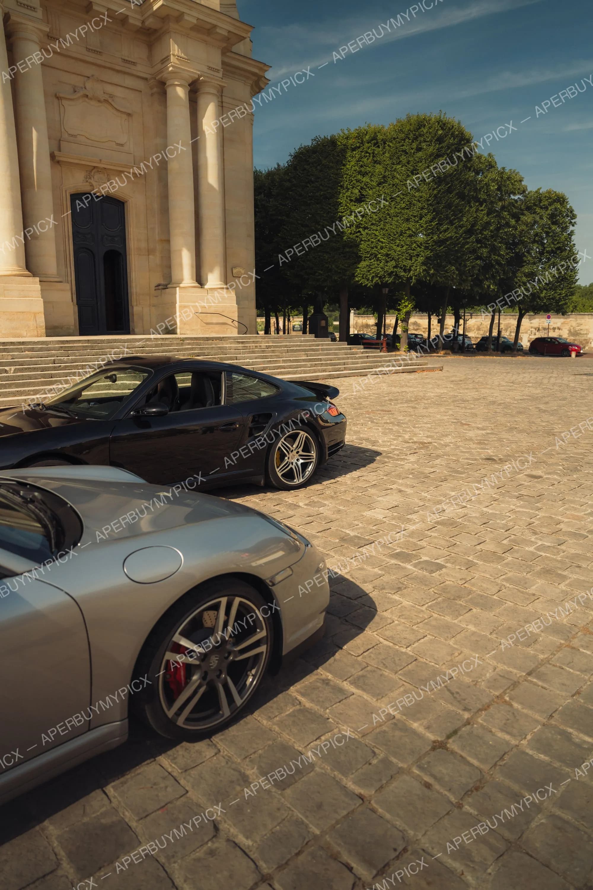 Deux Porsche 911 Turbo, une noire et une grise, garées sur une place pavée devant une église en pierre, avec des arbres et un ciel bleu en arrière-plan par temps ensoleillé.