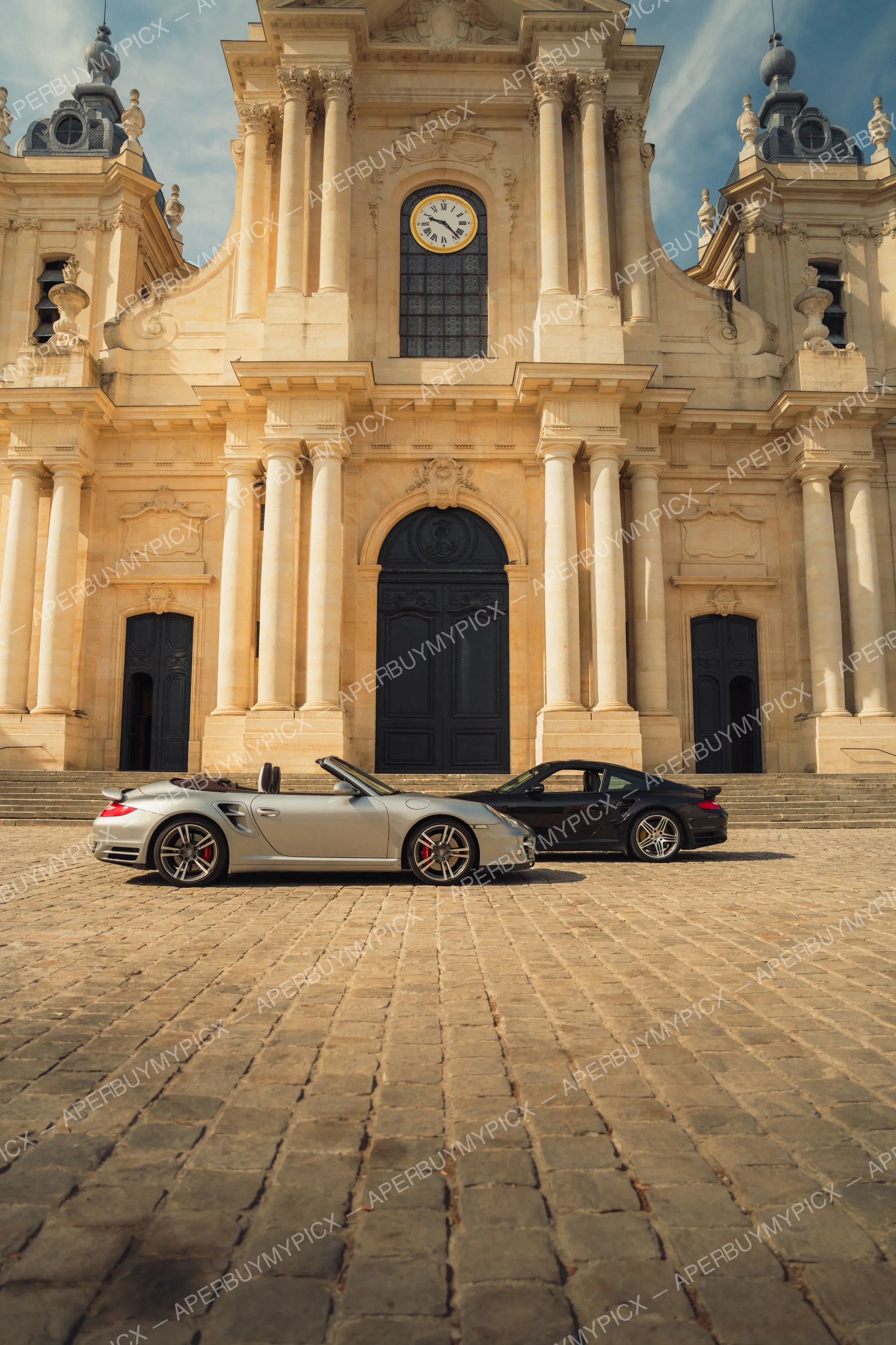 Une Porsche 911 Turbo cabriolet argentée et une Porsche 911 Turbo coupé noire garées sur une place pavée devant la façade baroque d'une cathédrale avec colonnes, horloge et grandes portes noires.