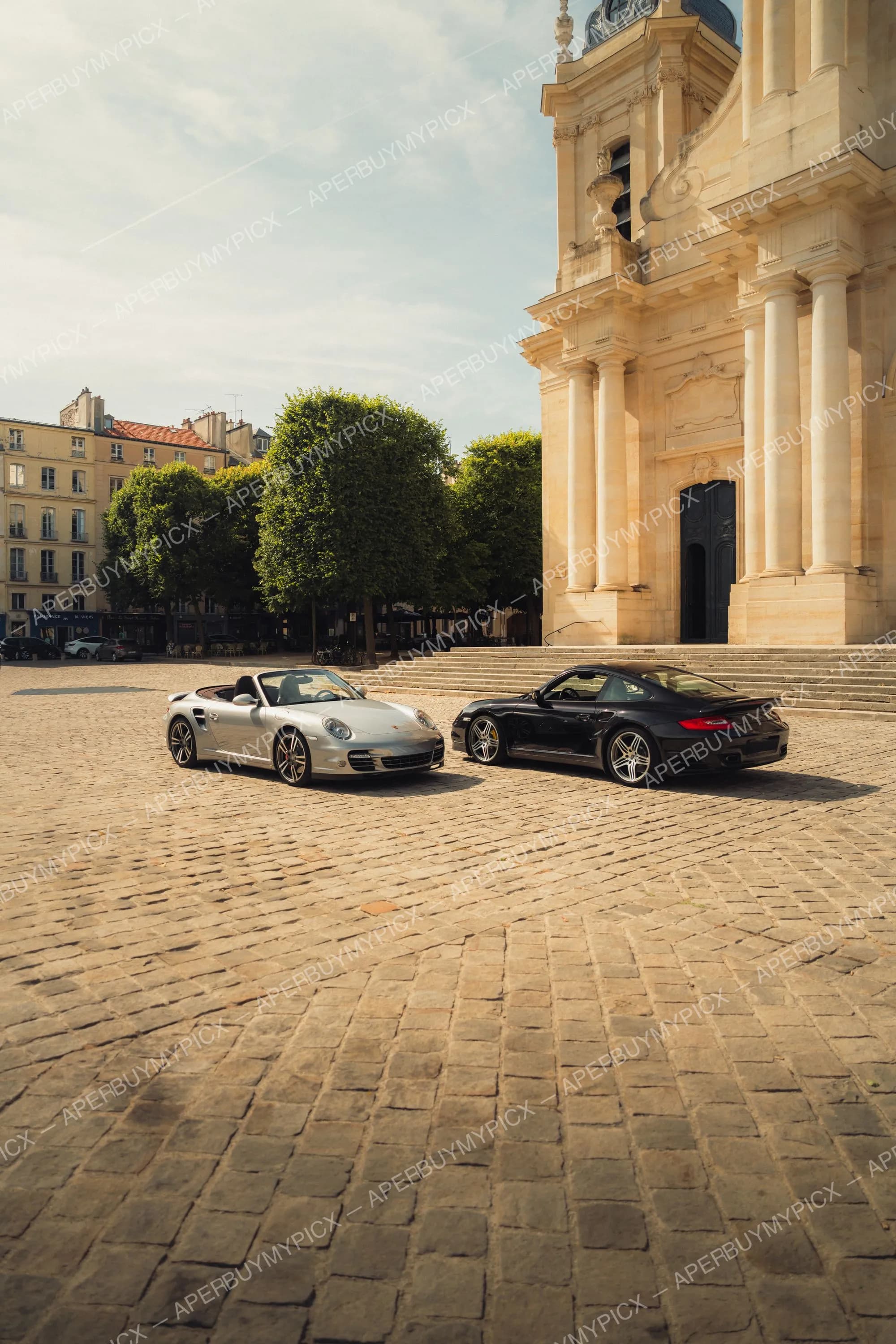 Deux Porsche 911 Turbo, une cabriolet argentée et un coupé noir, stationnées sur une place pavée devant une église classique en pierre à Paris, sous un ciel ensoleillé.