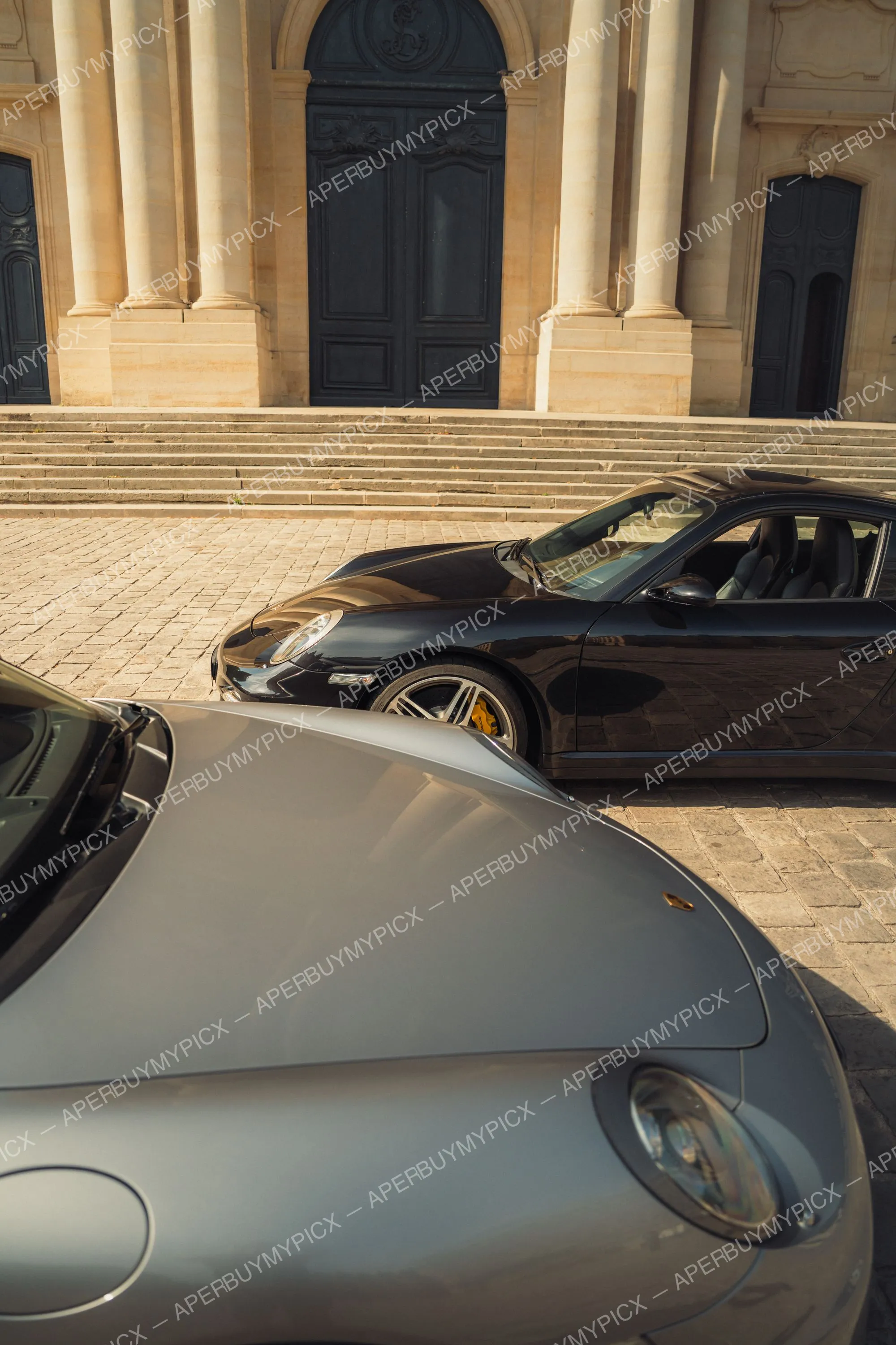 Porsche 911 Turbo noire aux étriers jaunes garée sur pavés devant un bâtiment classique à colonnes et portes noires, avec l'avant d'une Porsche grise au premier plan.