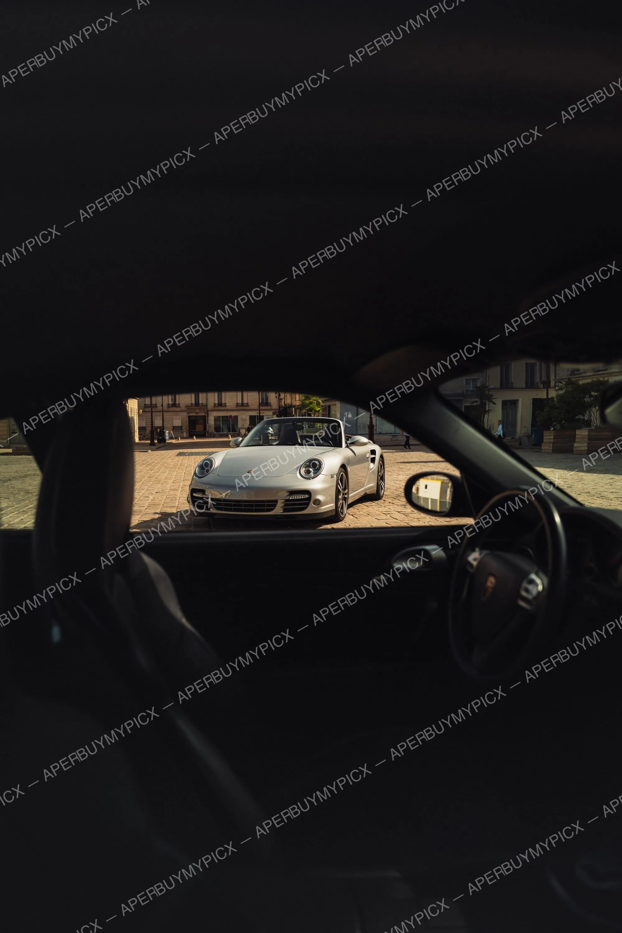Porsche 911 Turbo Cabriolet argentée garée sur une place pavée parisienne, vue à travers la vitre latérale d'une autre voiture au coucher du soleil