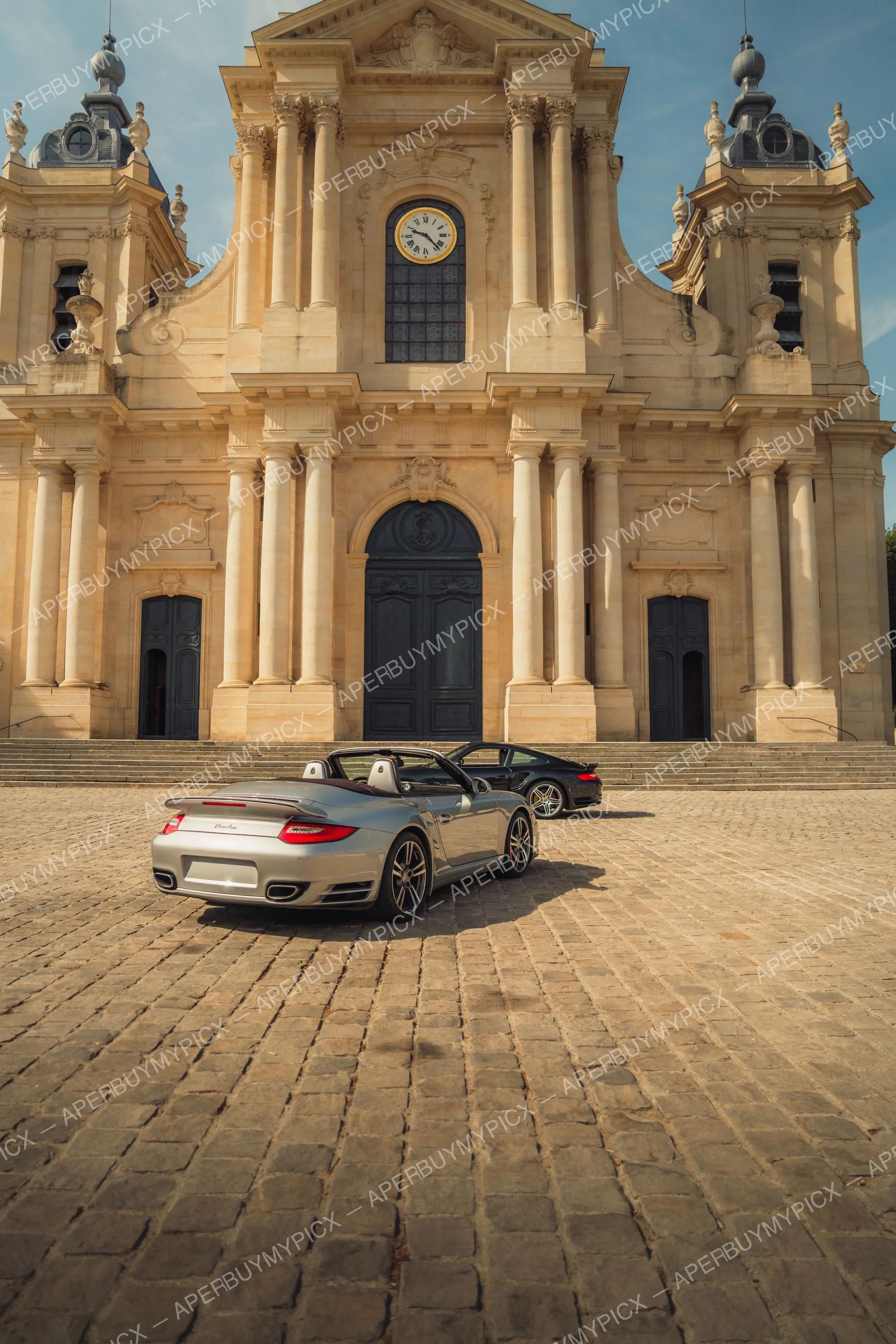 Deux Porsche 911 Turbo, un cabriolet argenté et un coupé noir, garées sur une place pavée devant la façade baroque d'une cathédrale avec horloge sous un ciel bleu.
