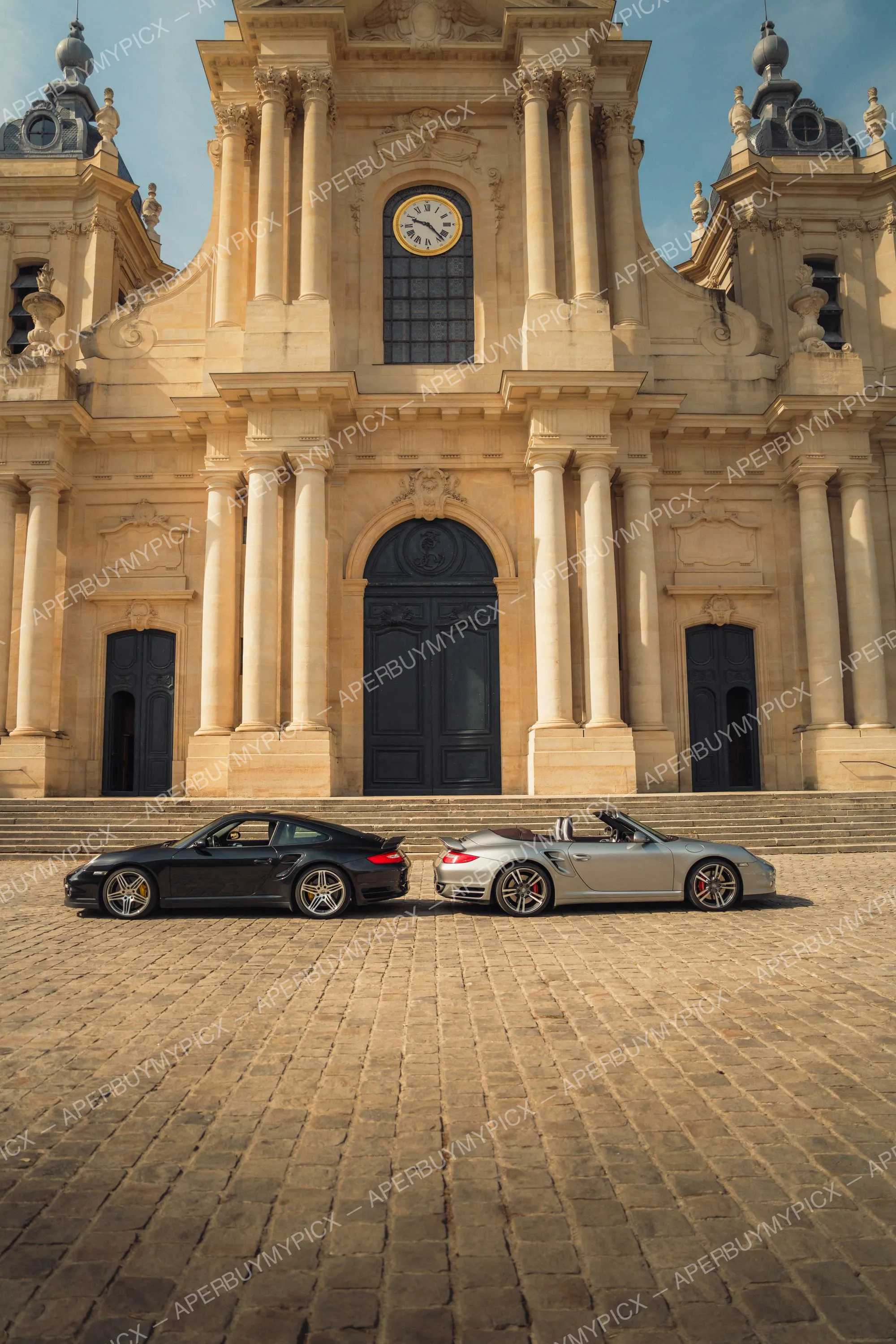 Une Porsche 911 Turbo coupé noire et une Porsche 911 Turbo cabriolet argent garées face à face sur un pavé devant la façade baroque d'une cathédrale avec horloge dorée.