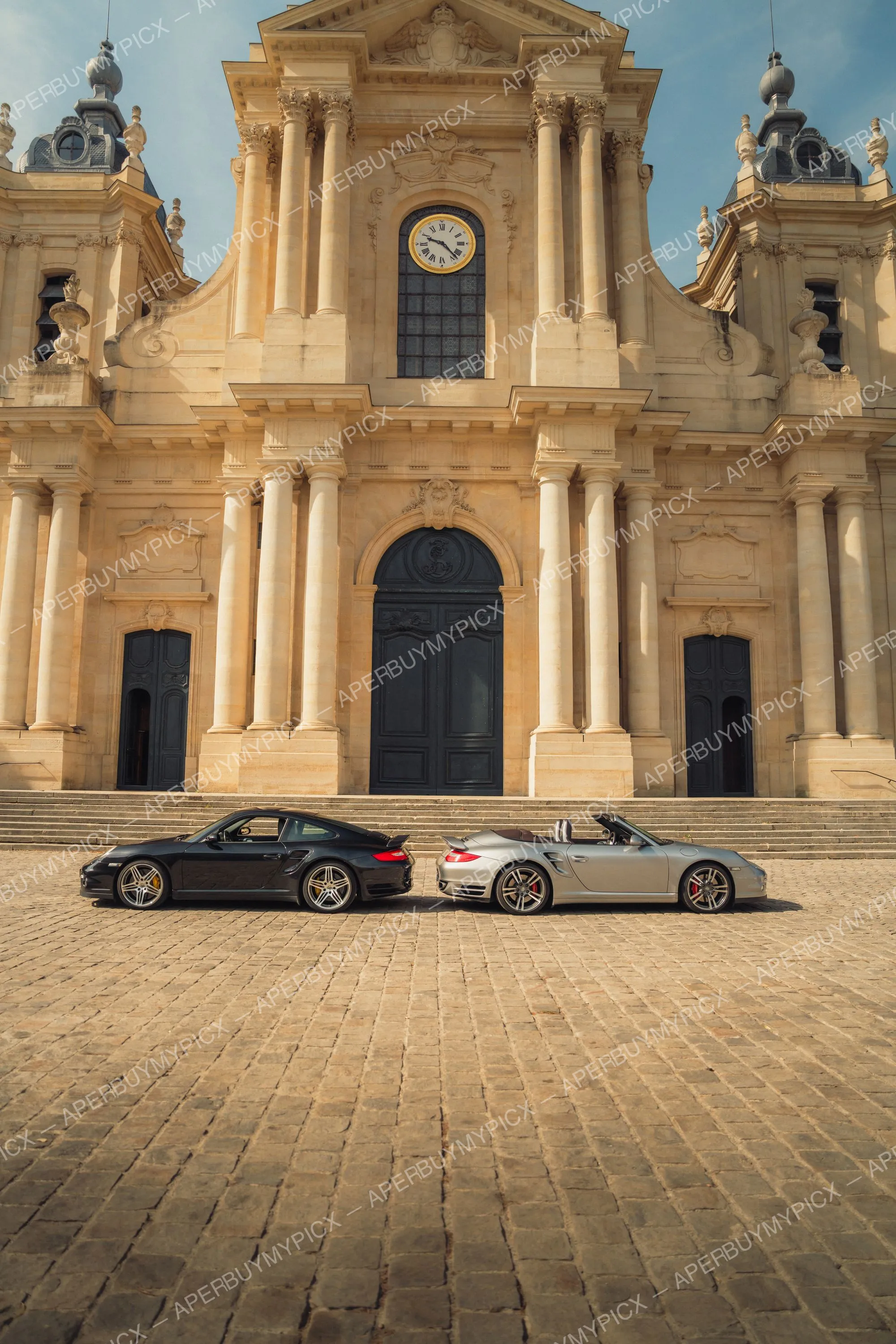 Deux Porsche 911 Turbo, une coupé noire et une cabriolet argentée, garées face à face sur des pavés devant une imposante église baroque à horloge et colonnes.