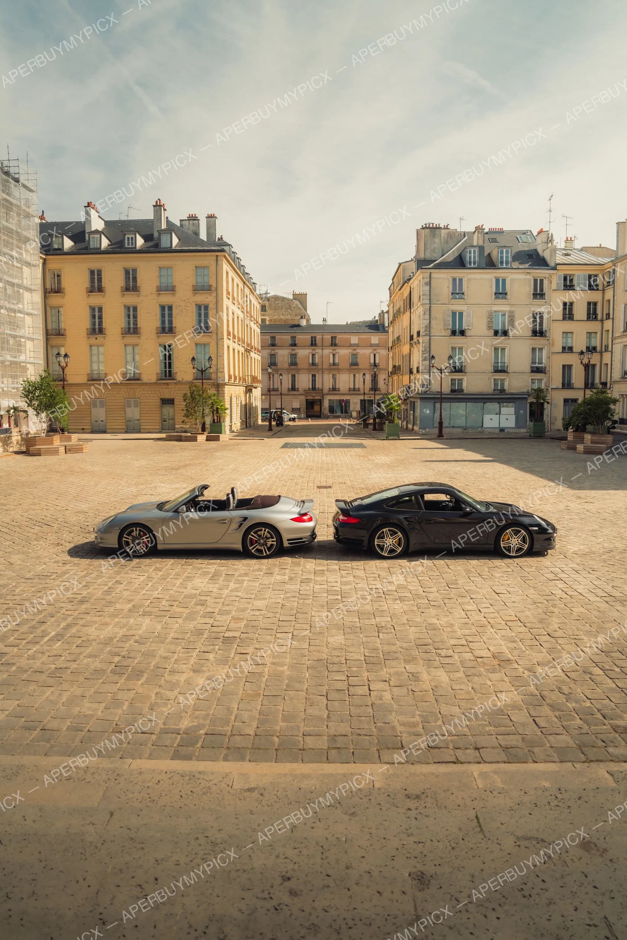 Une Porsche 911 Turbo cabriolet argentée et une Porsche 911 Turbo coupé noire garées nez à nez sur une place pavée parisienne bordée d'immeubles haussmanniens.
