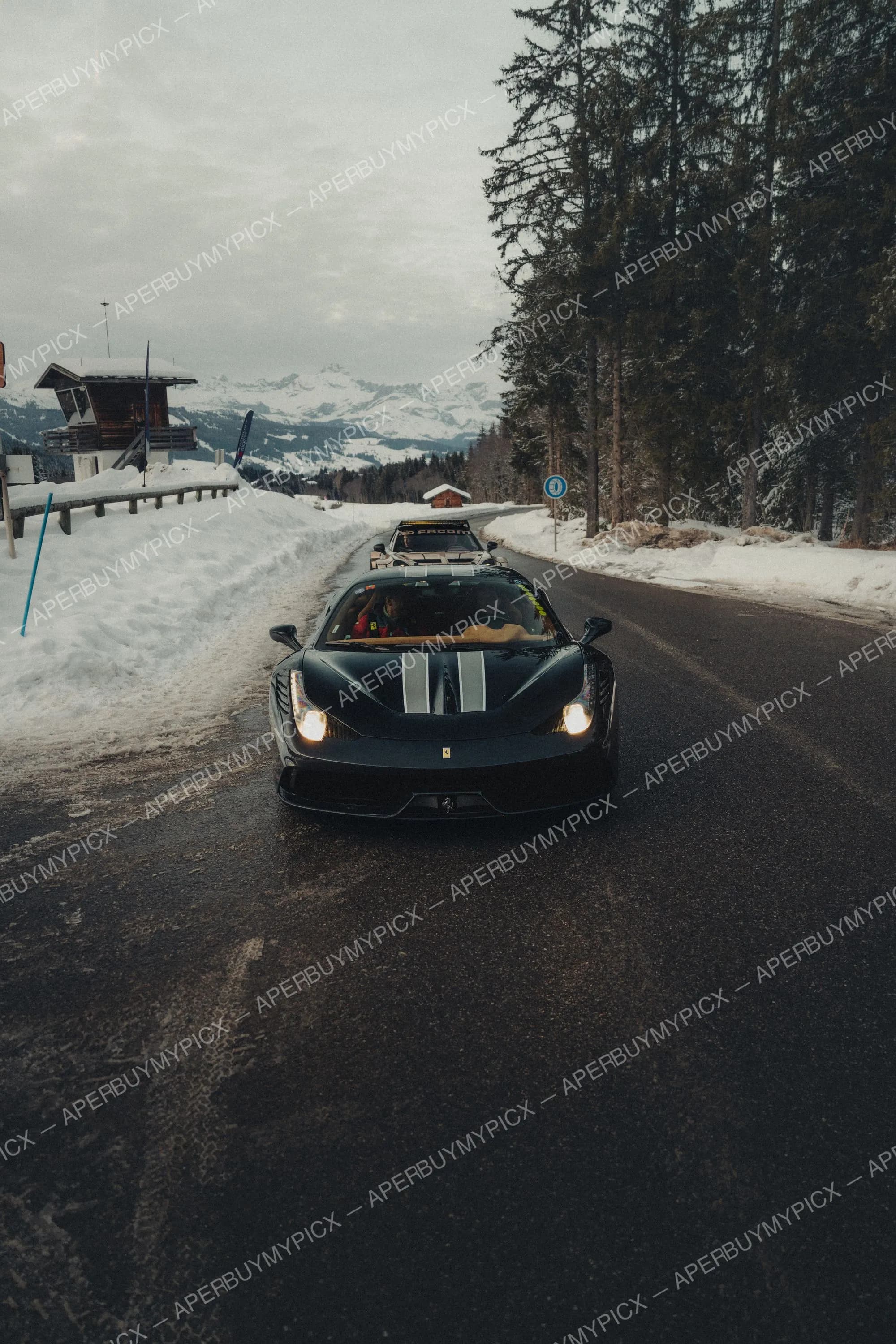 Ferrari 458 Speciale noire avec bandes grises roulant sur une route de montagne enneigée à Megève, phares allumés, sommets alpins et sapins en arrière-plan sous un ciel gris.