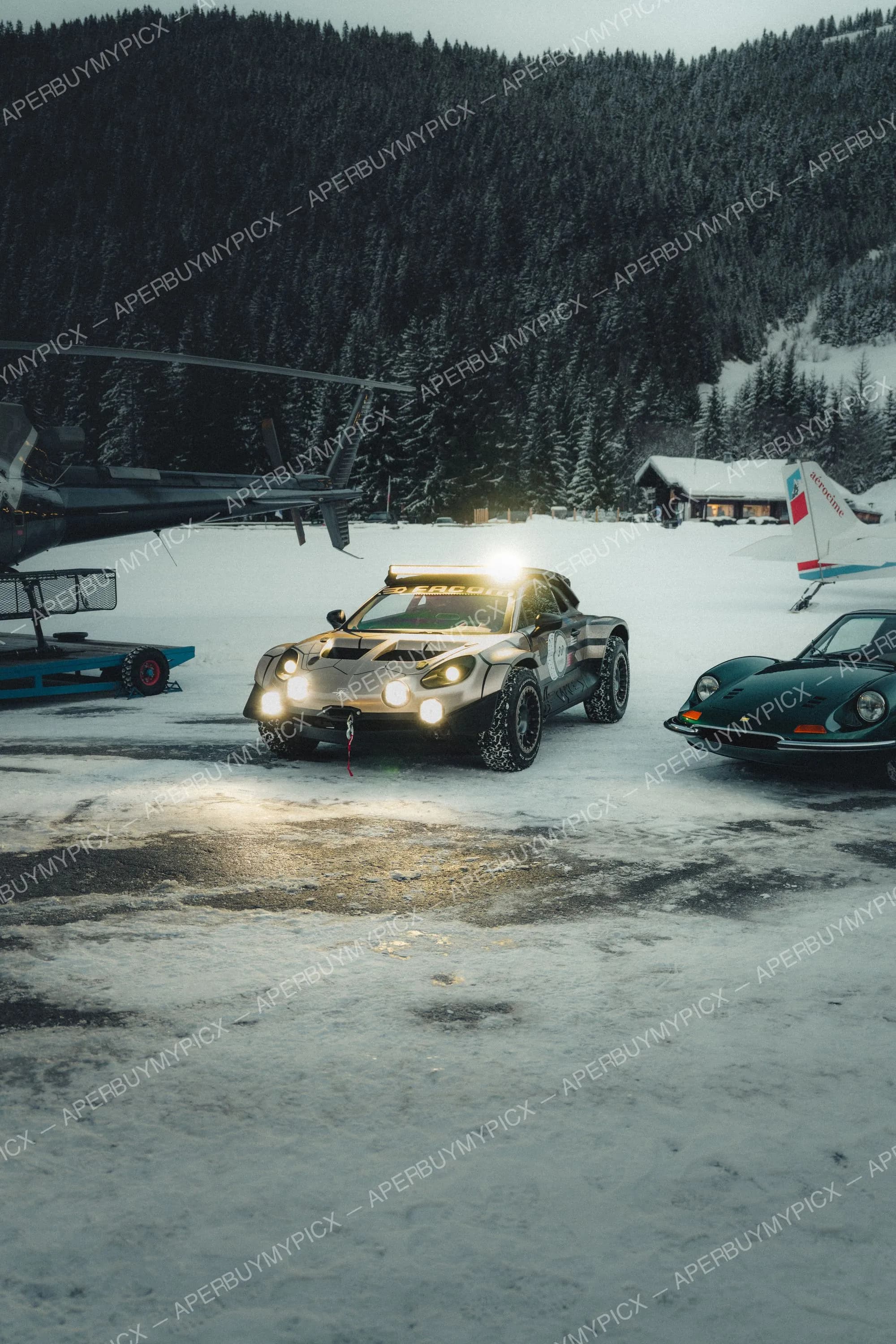 Voiture de rallye raid tout-terrain aux phares allumés stationnée dans la neige à Megève, entourée d'un hélicoptère, d'une Ferrari Dino verte et d'un avion, devant une forêt de sapins enneigée au crépuscule.