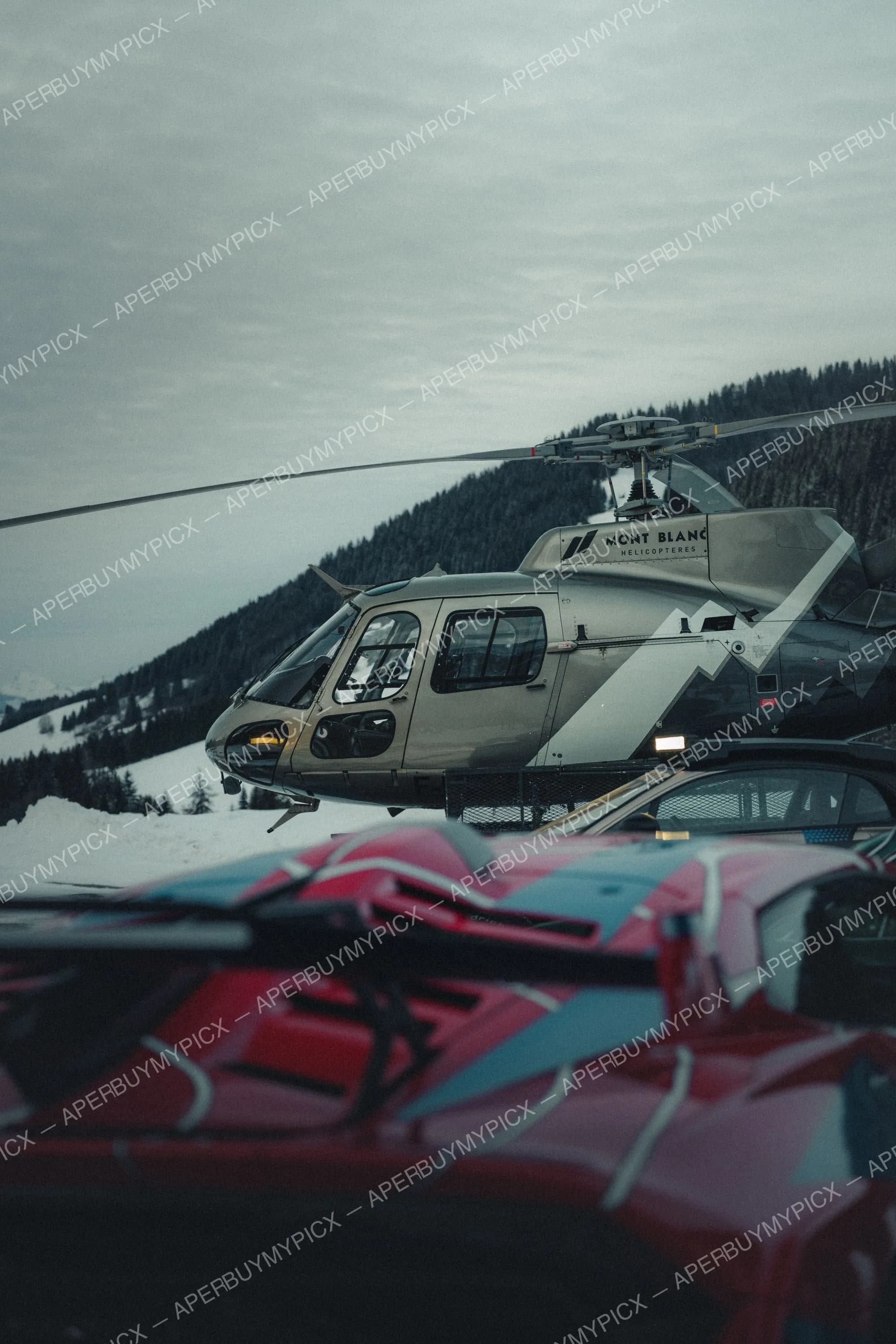 Hélicoptère Mont Blanc Hélicoptères stationné sur la neige à Megève, avec une supercar rouge et bleue au premier plan et montagnes boisées enneigées en arrière-plan sous un ciel gris.