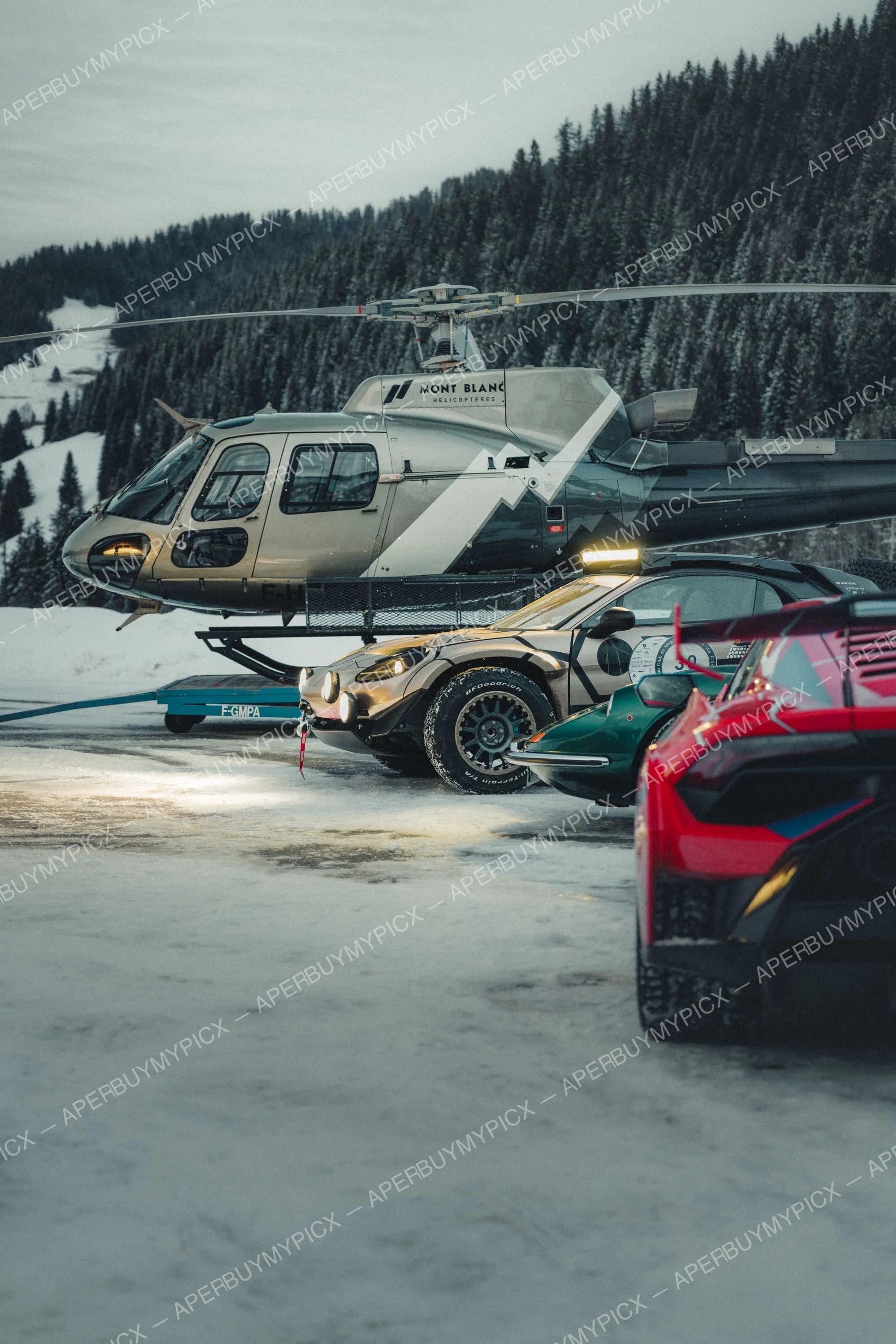 Hélicoptère Mont Blanc Hélicoptères et voitures de rallye dorée, verte et rouge alignées sur la neige devant une montagne boisée à Megève