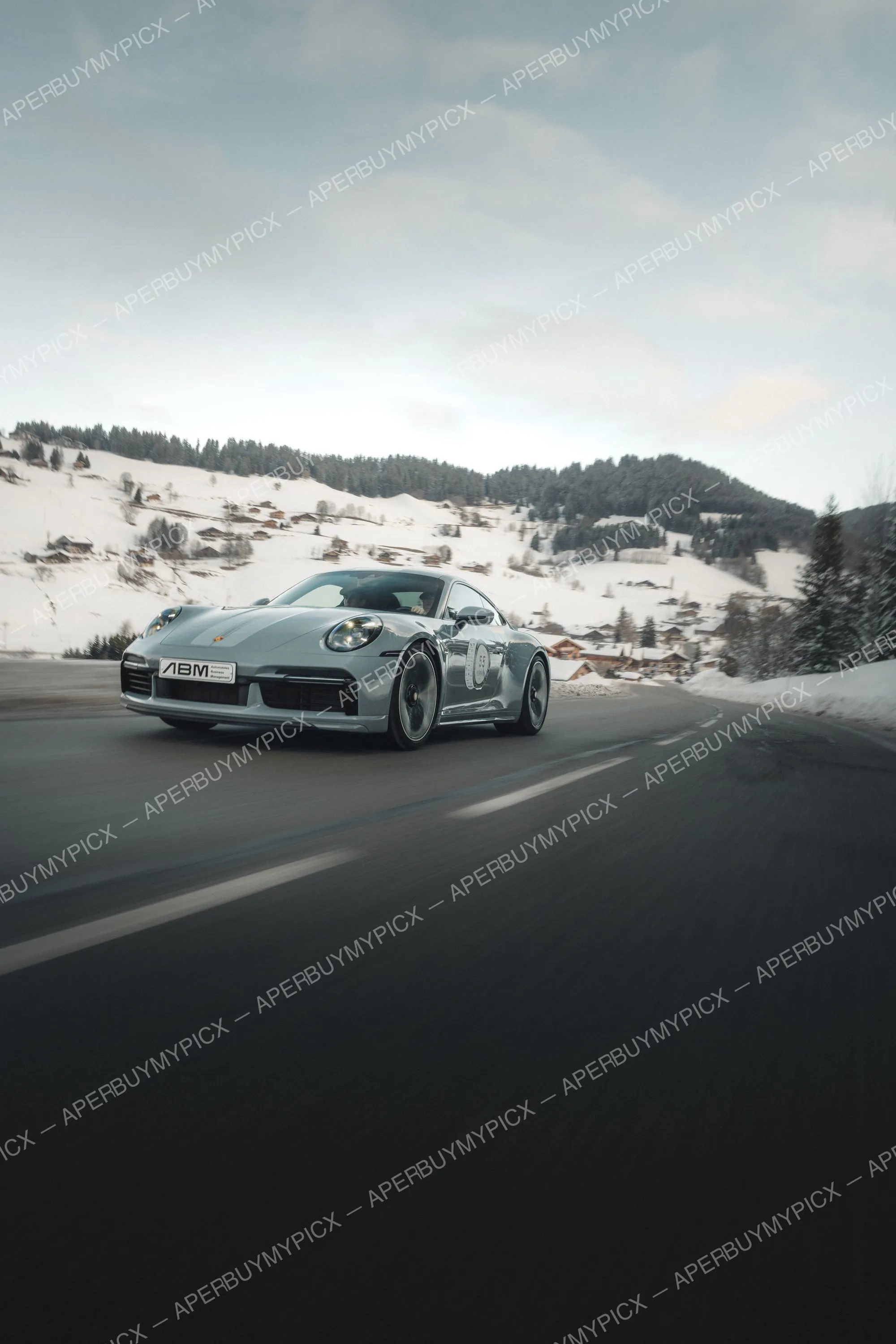 Porsche 911 grise roulant sur une route de montagne bordée de neige, avec un village alpin enneigé et des sapins en arrière-plan à Megève.