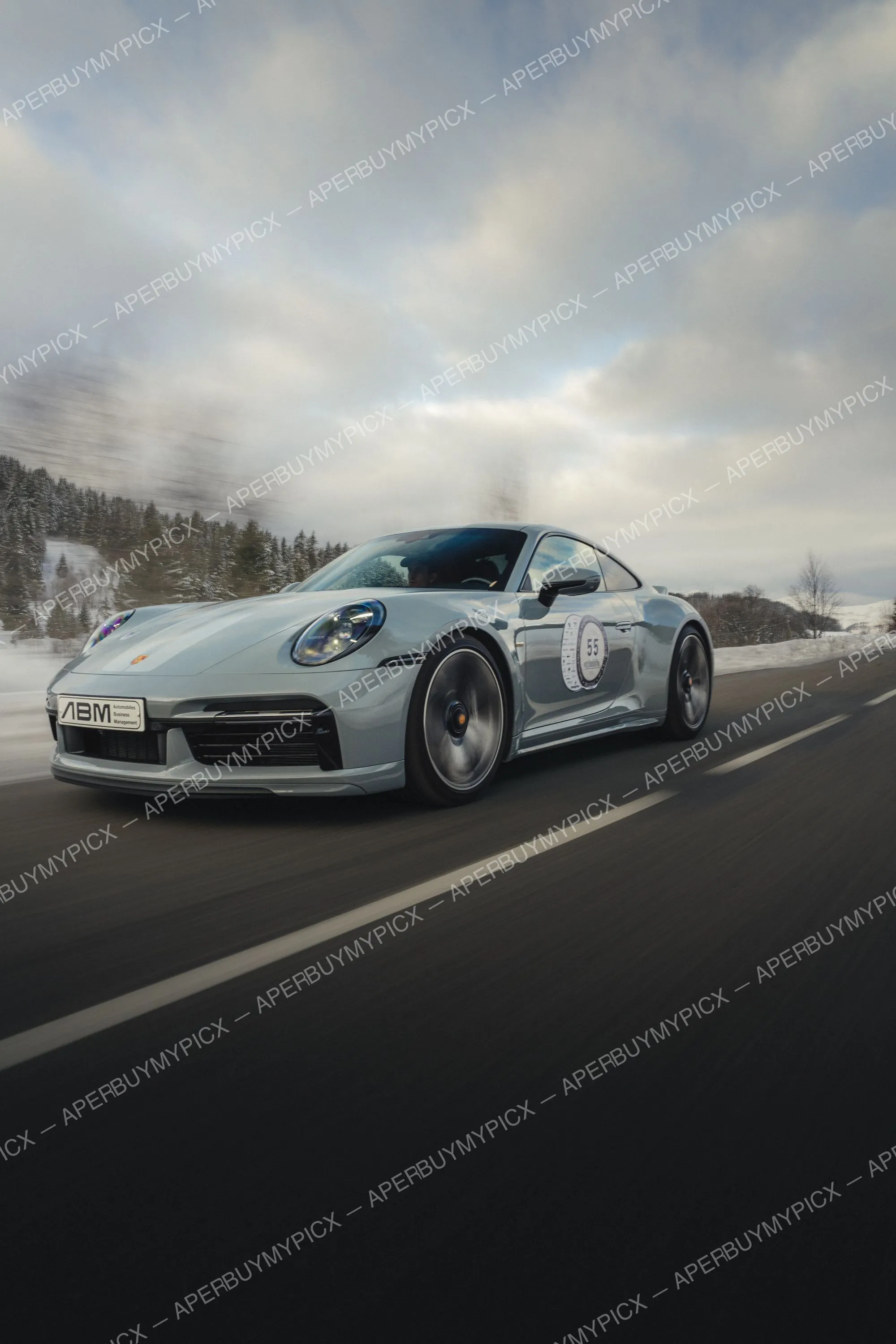 Porsche 911 Sport Classic grise numéro 55 roulant sur une route de montagne enneigée près de Megève sous un ciel nuageux