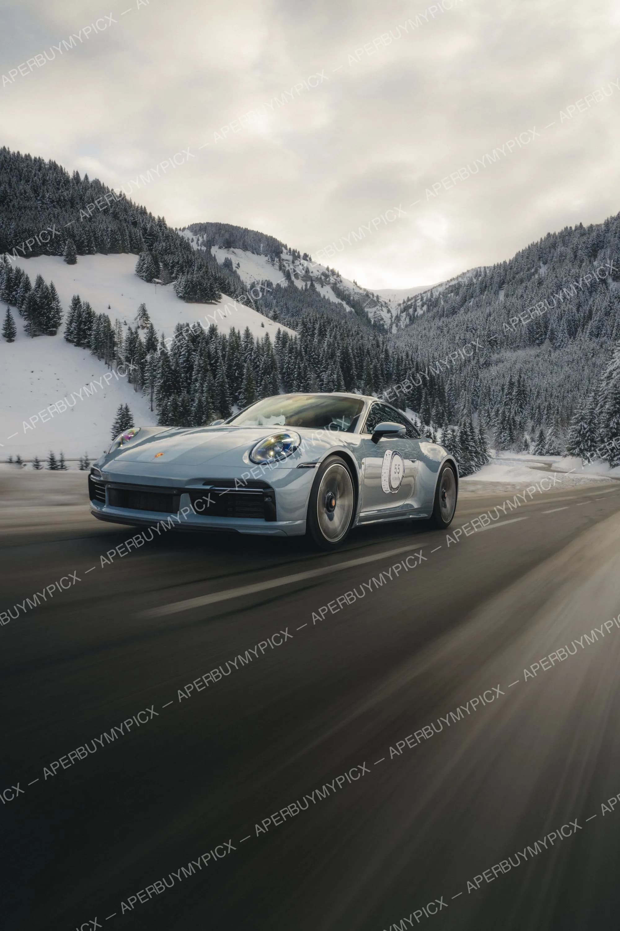 Porsche 911 bleu gris en mouvement sur une route de montagne enneigée près de Megève, avec forêts de sapins et sommets alpins enneigés en arrière-plan sous un ciel nuageux.