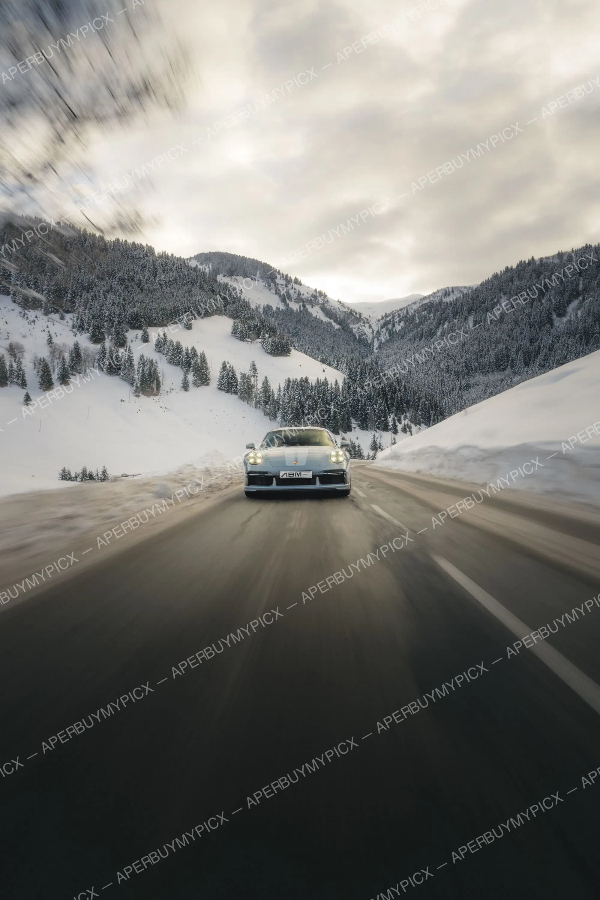 Porsche 911 grise roulant sur une route de montagne bordée de sapins enneigés sous un ciel nuageux, avec effet de vitesse.