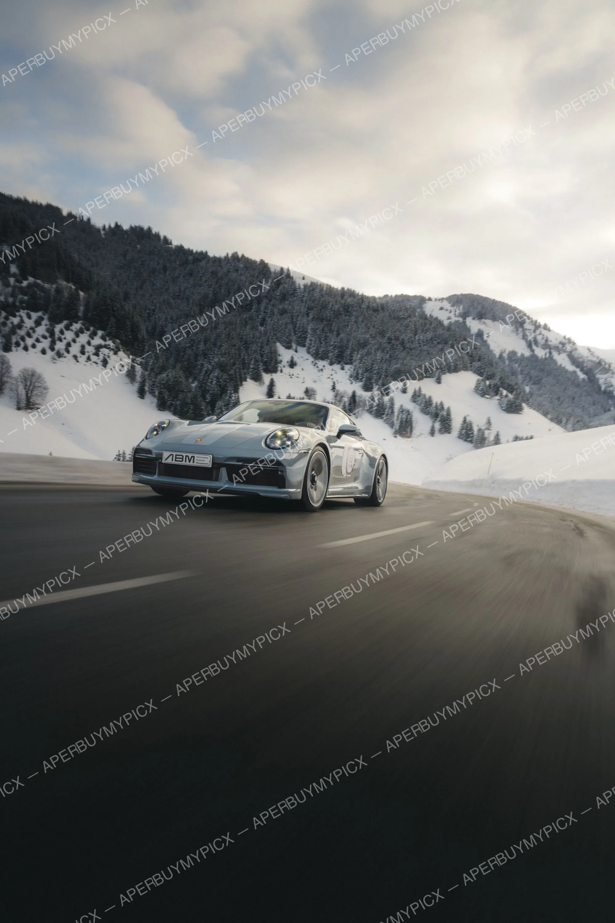 Porsche 911 grise roulant sur une route de montagne enneigée à Megève, entourée de sapins et sommets alpins sous un ciel nuageux.