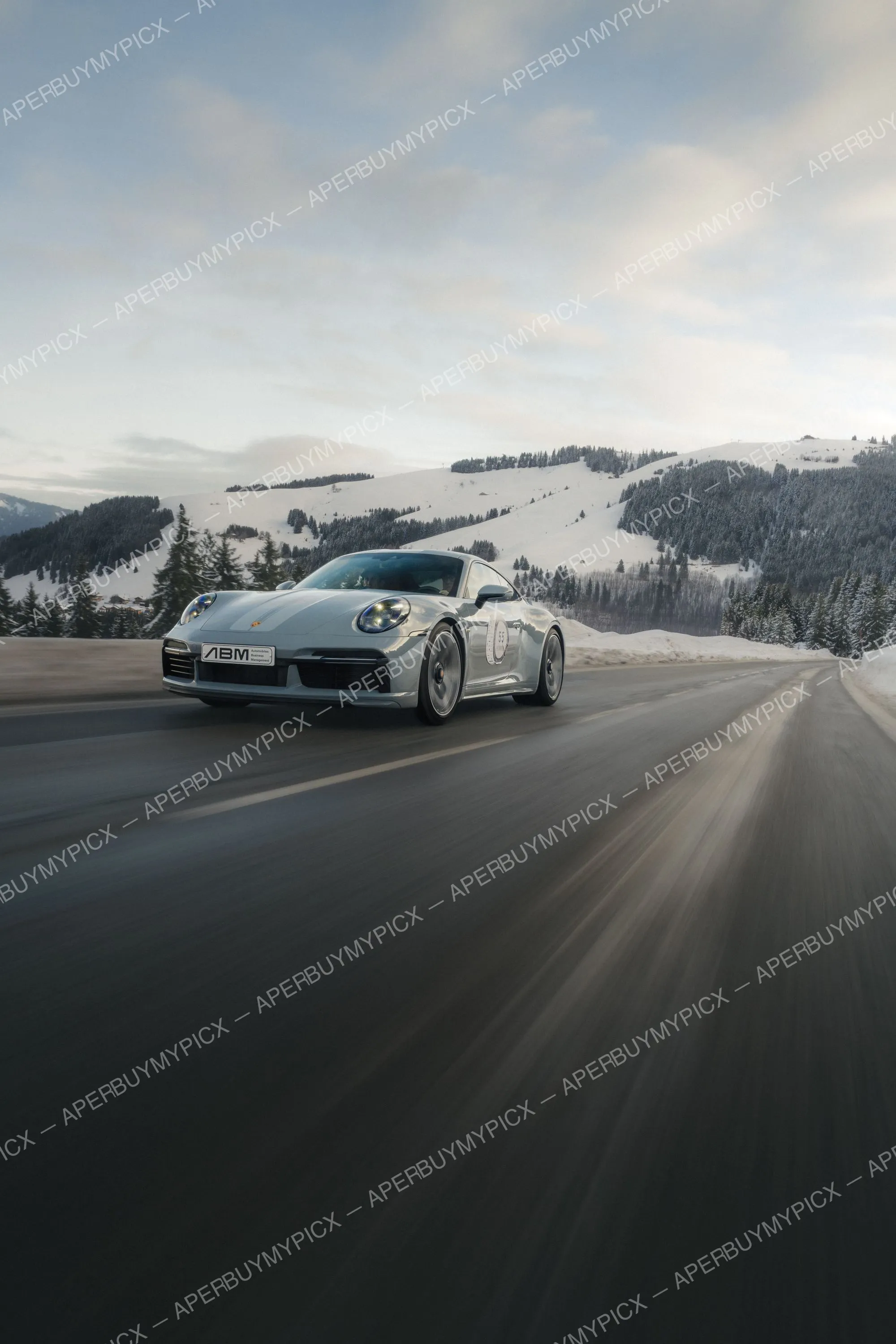 Porsche 911 Sport Classic grise roulant sur une route de montagne bordée de sapins et de pentes enneigées à Megève sous un ciel nuageux