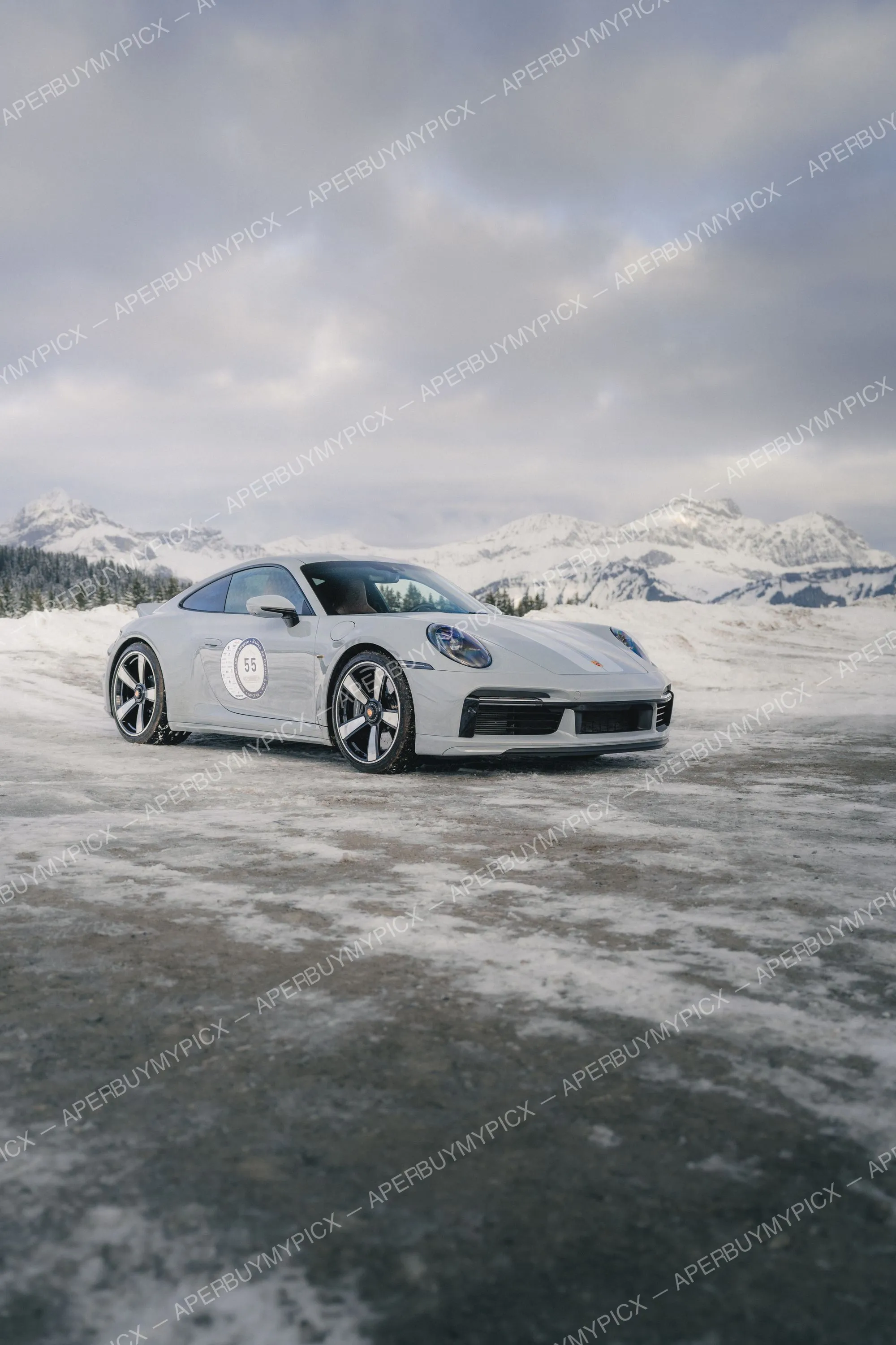 Porsche 911 blanche portant le numéro 55 stationnée sur une route enneigée devant des montagnes alpines sous un ciel nuageux à Megève