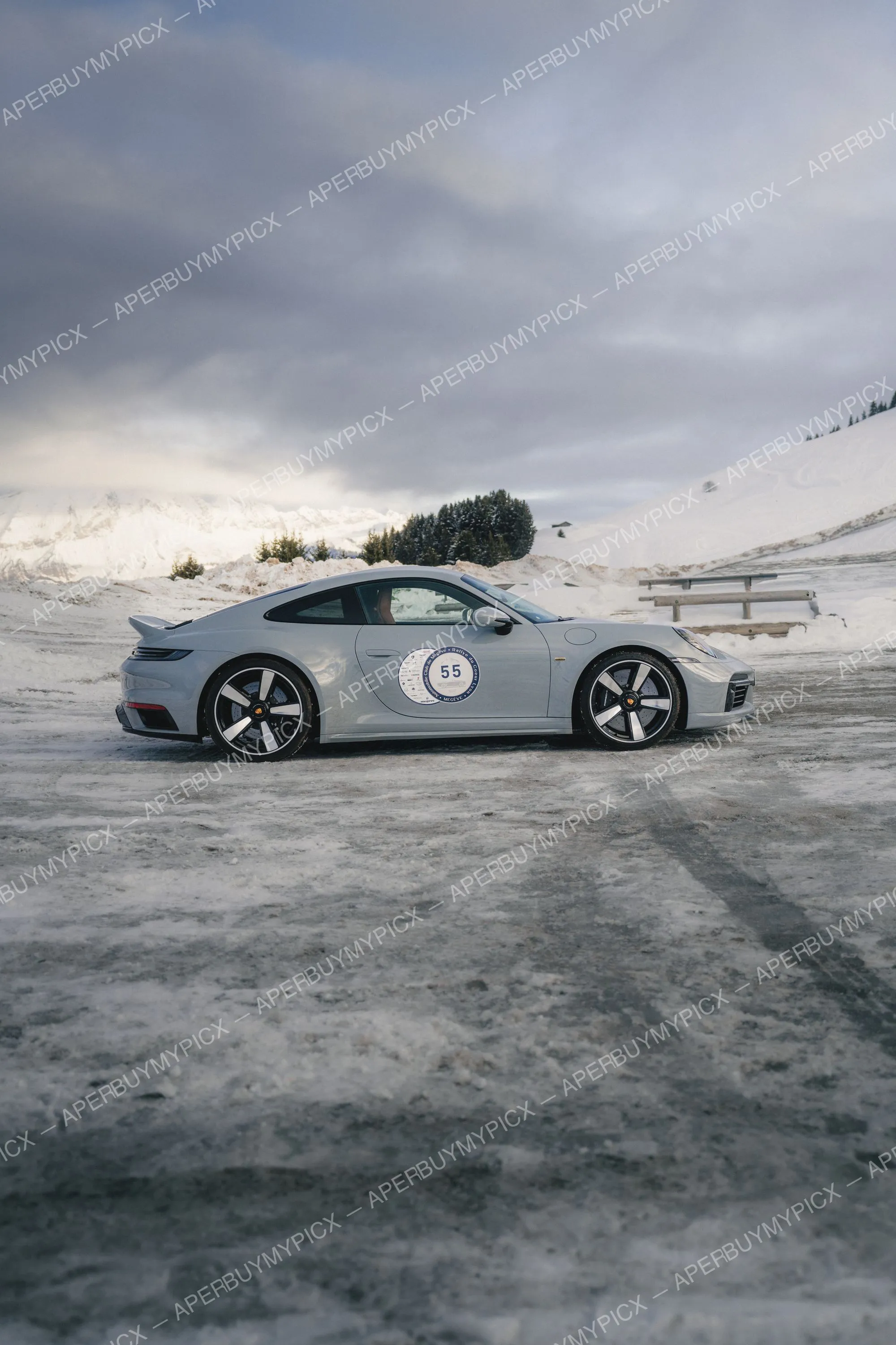 Porsche 911 grise numéro 55 du Rallye du Cœur de Megève garée sur un sol enneigé, montagnes et sapins enneigés en arrière-plan sous un ciel nuageux.