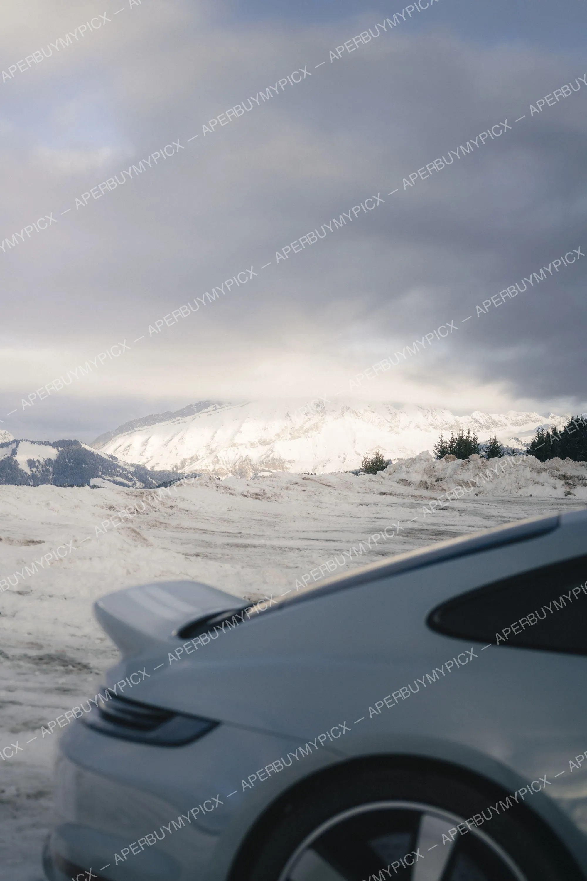 Arrière d'une voiture de sport blanche avec aileron, garée dans la neige devant un panorama de montagnes enneigées sous un ciel nuageux à Megève.
