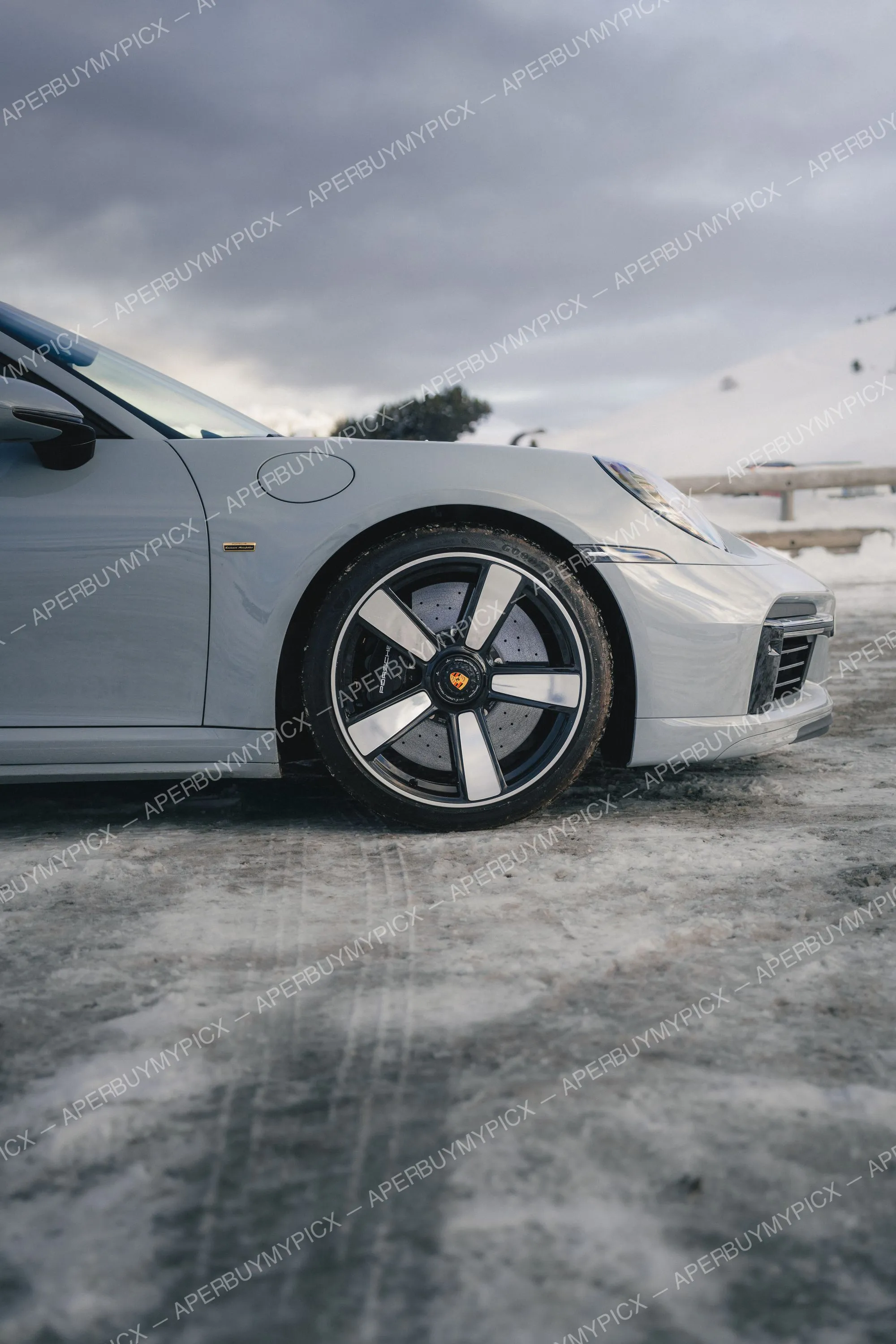 Avant d'une Porsche 911 grise garée sur une route enneigée à Megève, avec jantes noires et argentées et ciel nuageux hivernal.