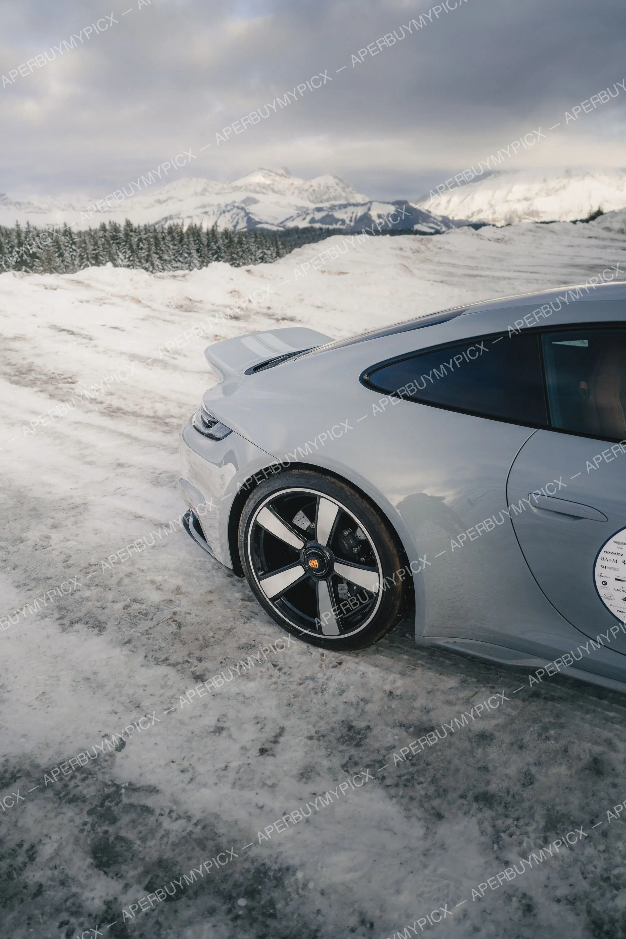 Porsche 911 grise vue arrière trois-quarts, jante noire et argent, stationnée sur la neige devant des montagnes enneigées et une forêt de sapins à Megève sous un ciel nuageux.