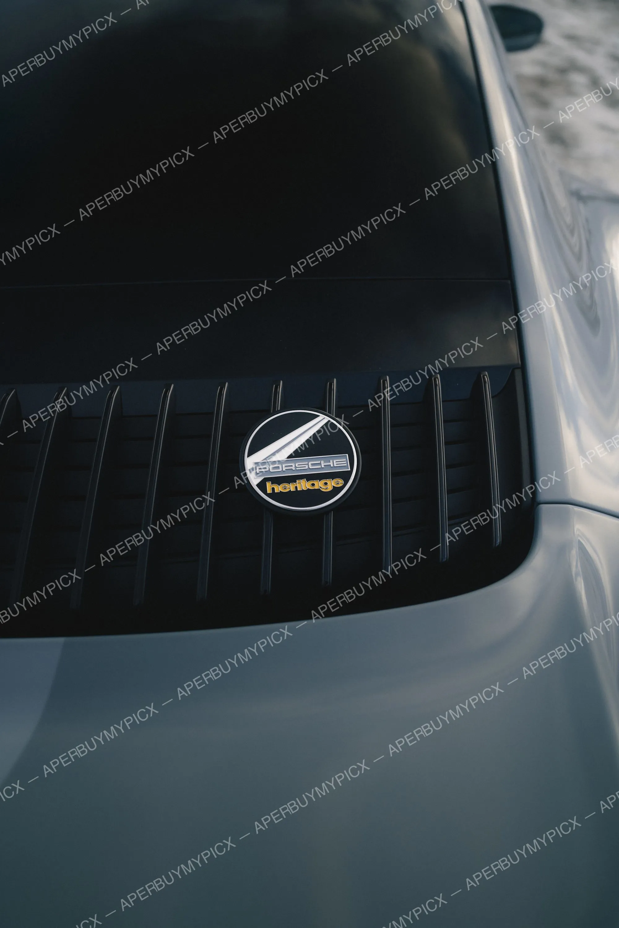 Gros plan du badge doré Porsche Heritage placé sur la grille moteur noire d'une Porsche argentée, capot en arrière-plan.
