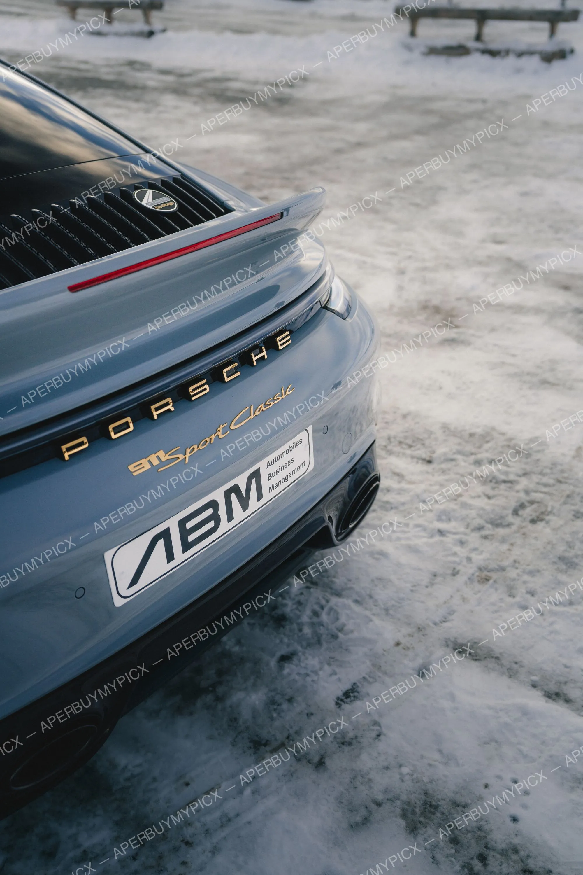 Vue arrière d'une Porsche 911 Sport Classic gris ardoise avec badge Porsche doré et aileron sur un parking enneigé à Megève