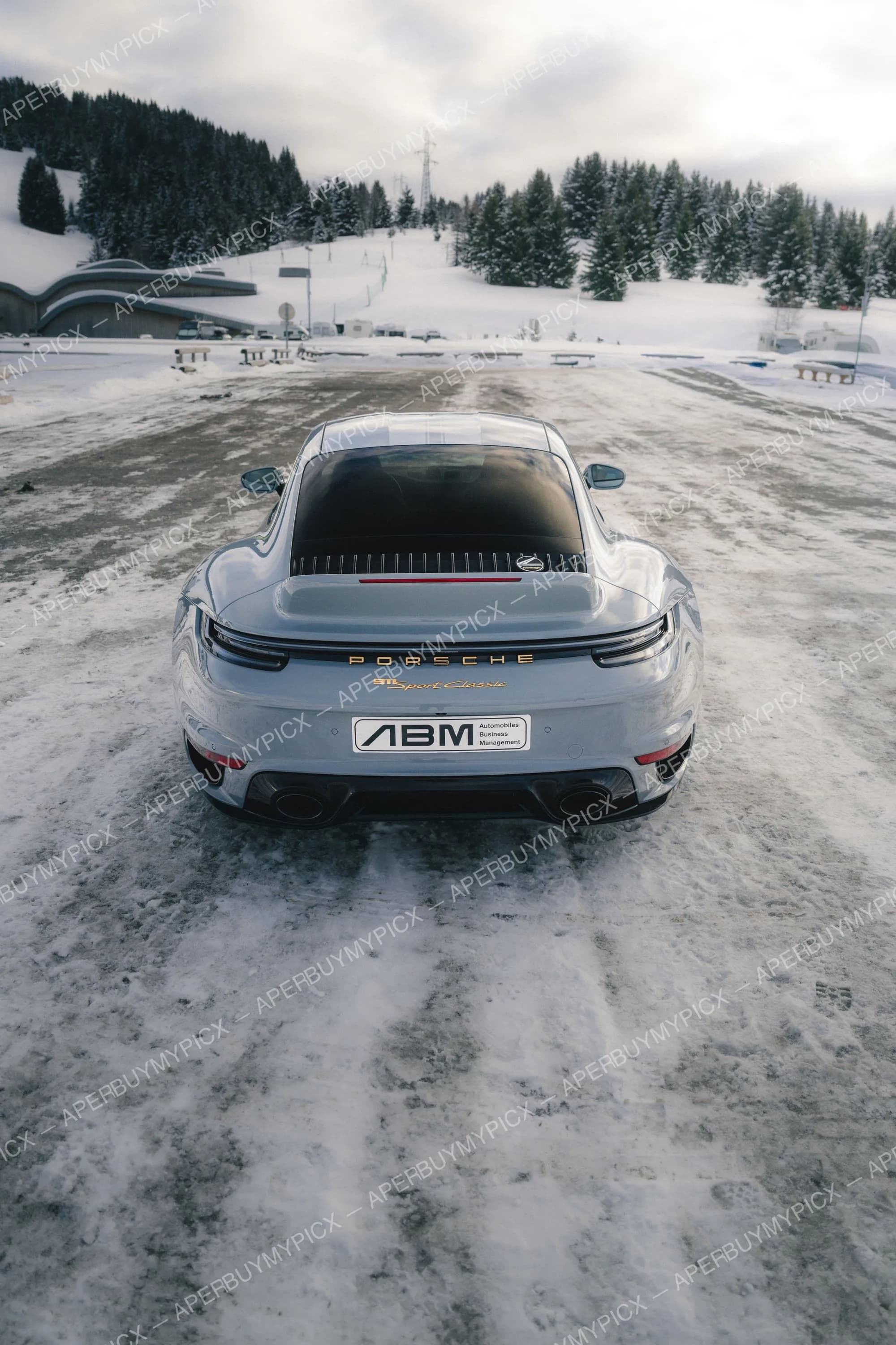 Vue arrière d'une Porsche 911 Sport Classic grise garée sur un sol enneigé à Megève, avec plaque ABM, montagnes boisées et pistes de ski en arrière-plan sous un ciel nuageux.