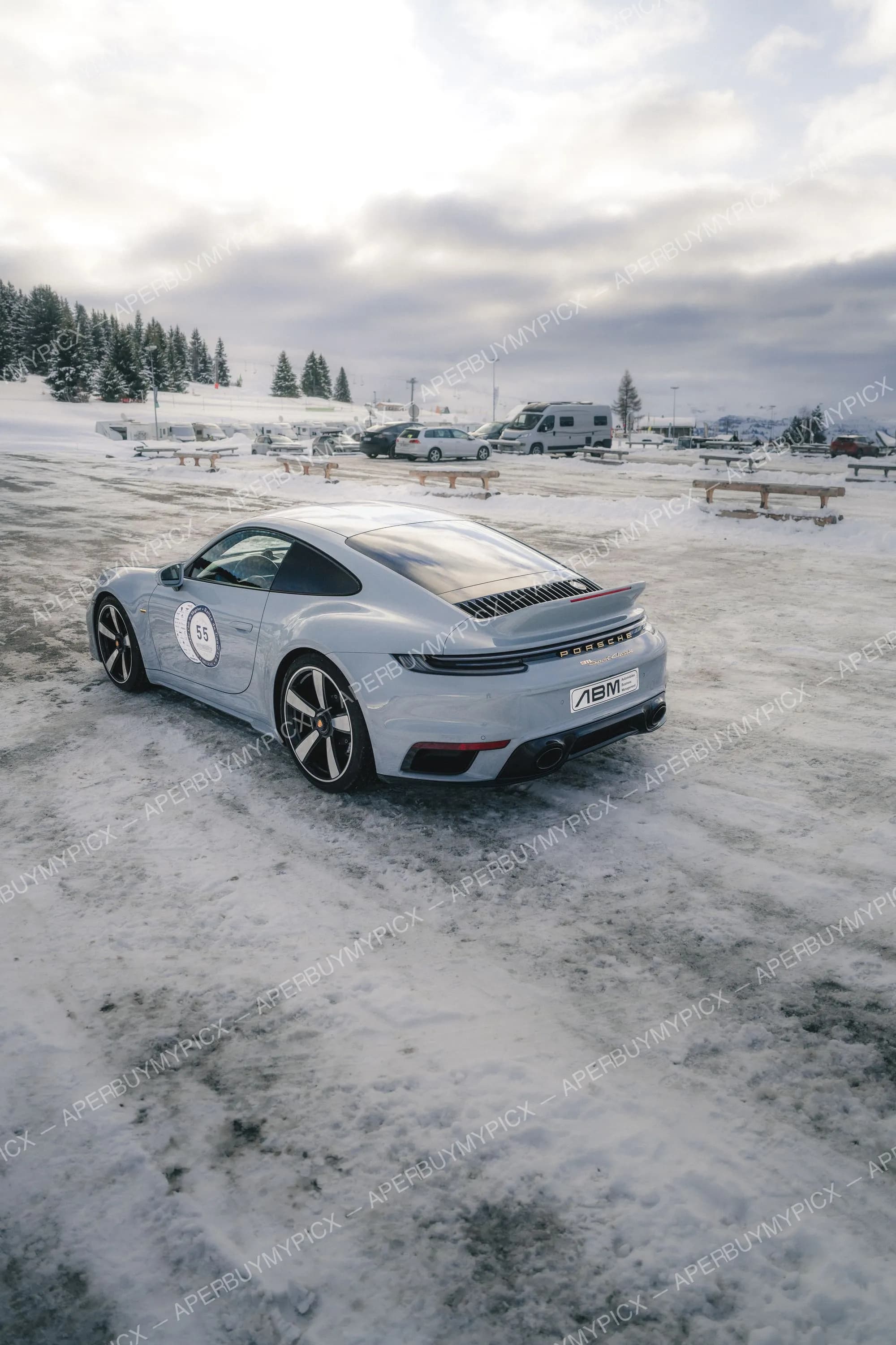 Porsche 911 Sport Classic grise portant le numéro 55 et le logo ABM, vue arrière trois-quarts sur un parking enneigé de station de ski à Megève, sous un ciel nuageux avec sapins et camping-cars en arrière-plan.