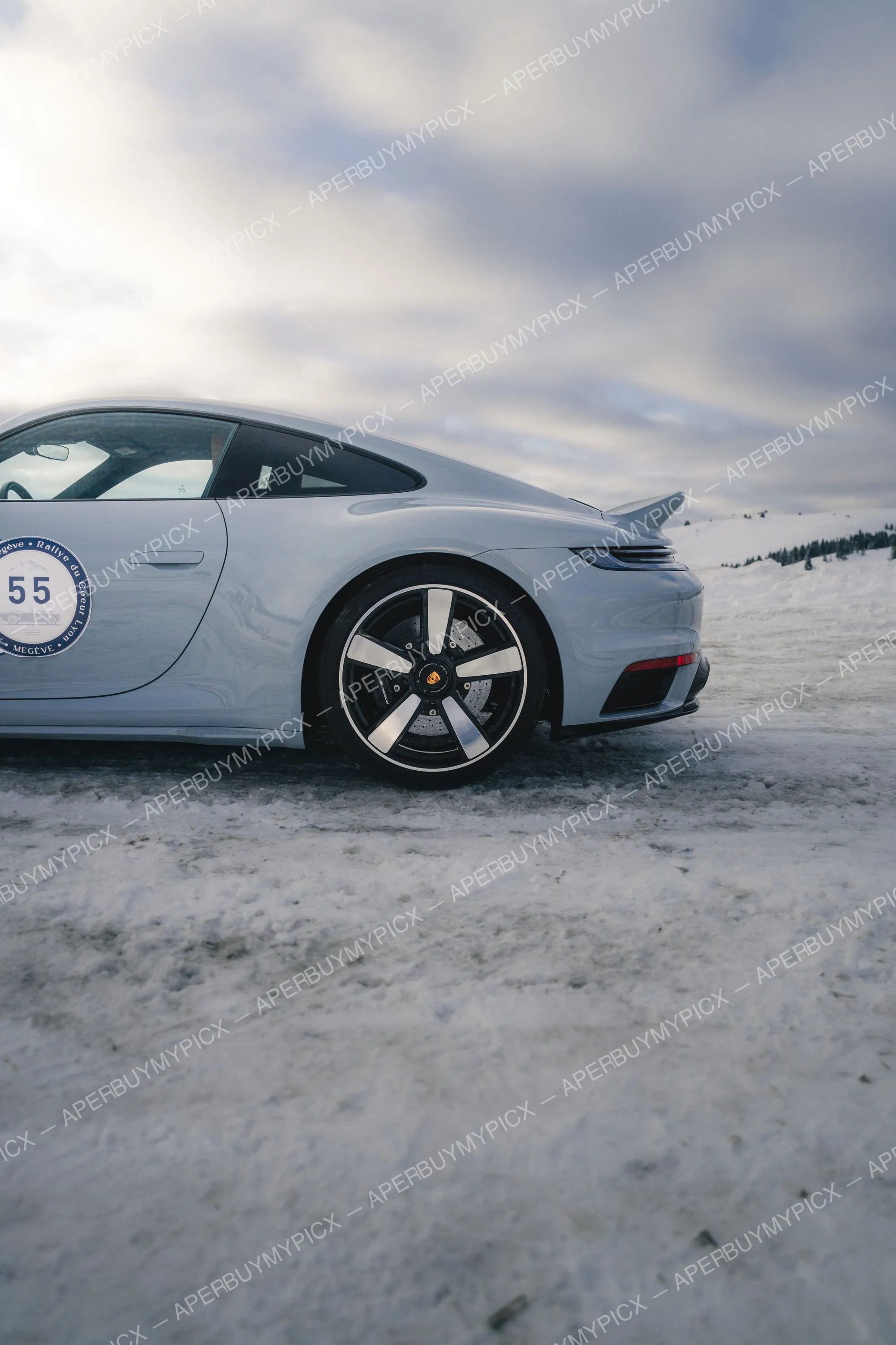 Porsche 911 grise vue de profil arrière portant le numéro 55 du Rallye du Cœur Lyon Megève, garée sur la neige sous un ciel nuageux en montagne