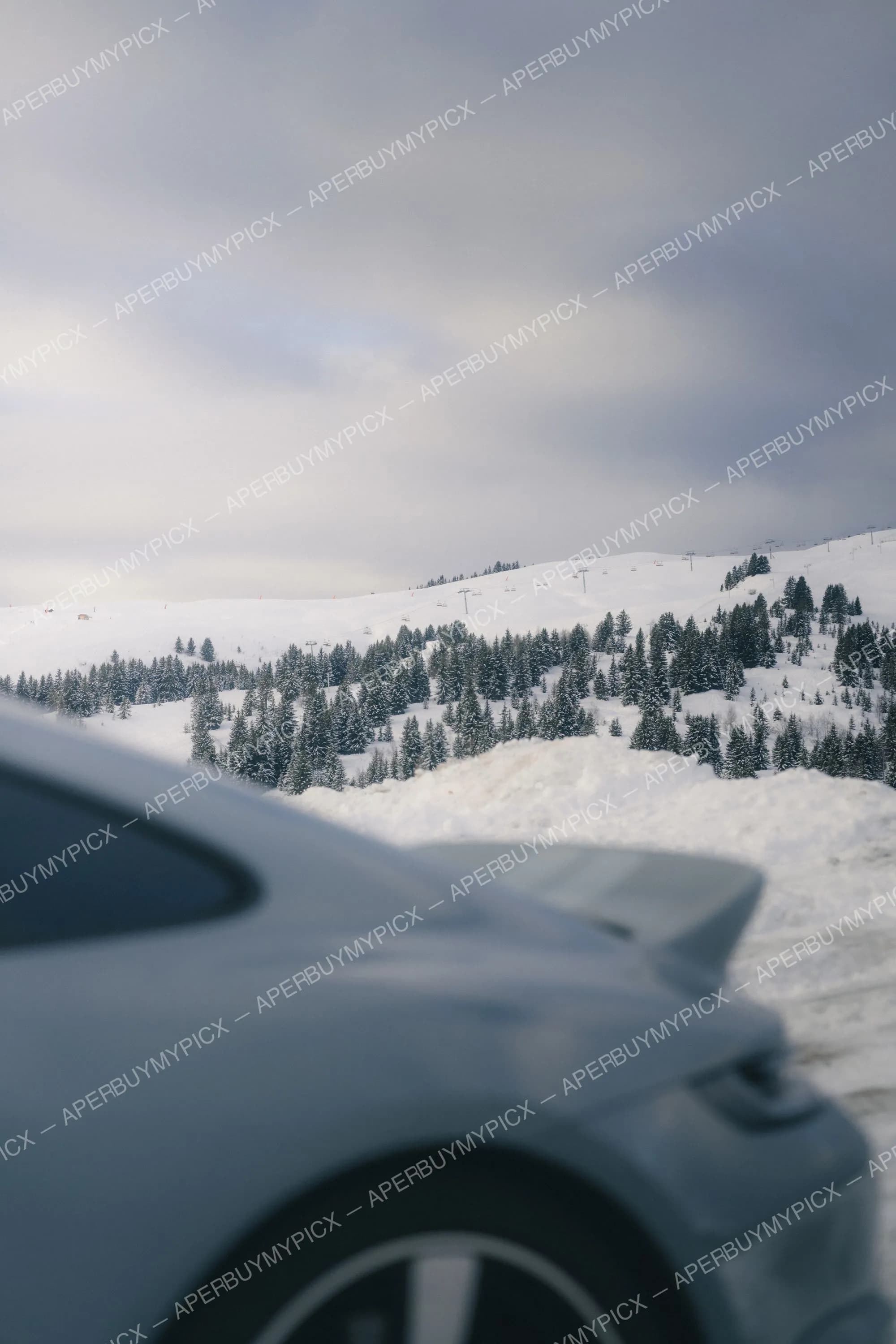 Carrosserie d'une voiture de sport grise devant un paysage alpin enneigé de Megève avec sapins, remontées mécaniques et ciel nuageux