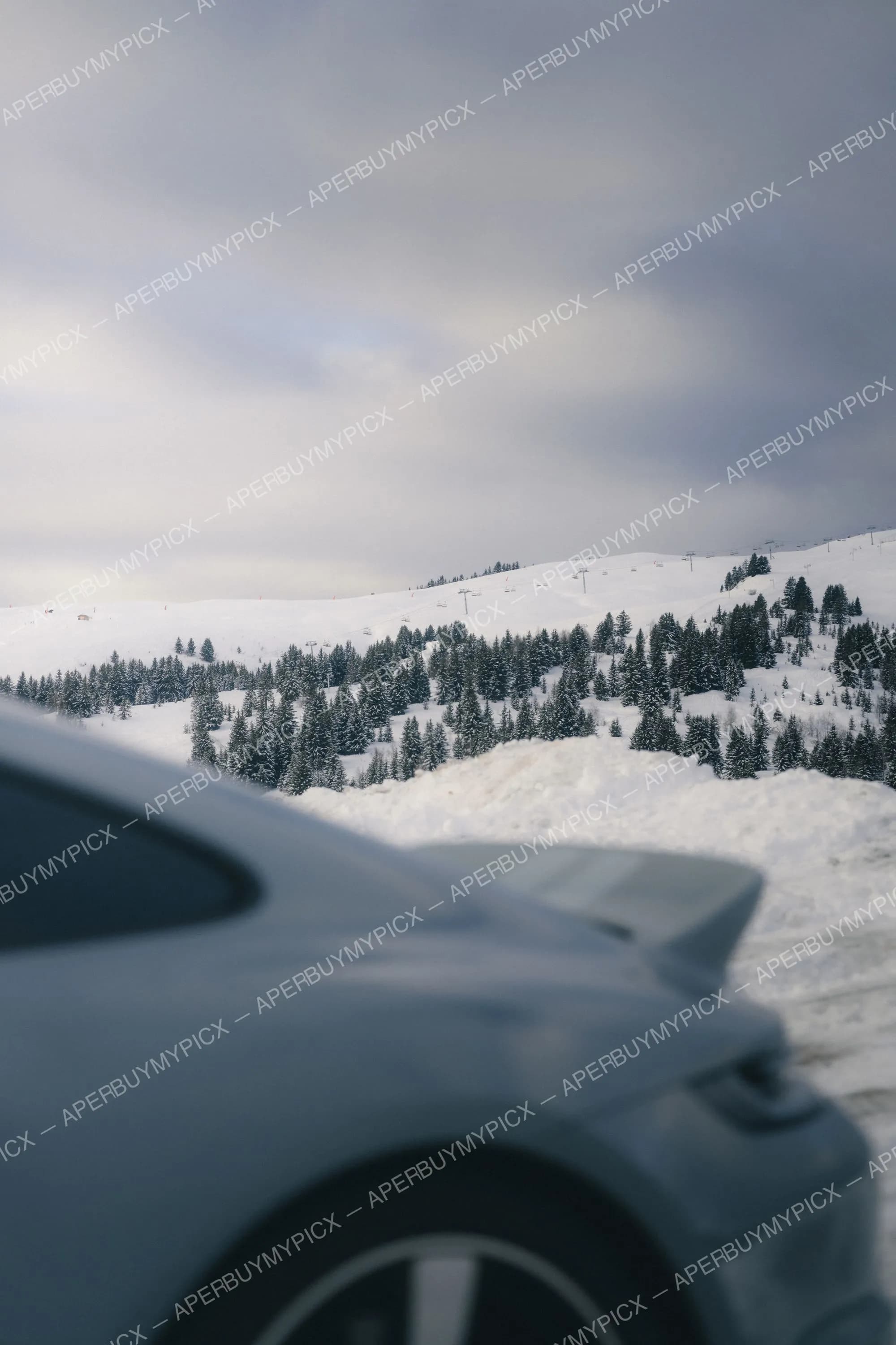 Aile avant d'une voiture de sport grise devant un paysage montagneux enneigé avec forêt de sapins et remontées mécaniques à Megève sous ciel nuageux