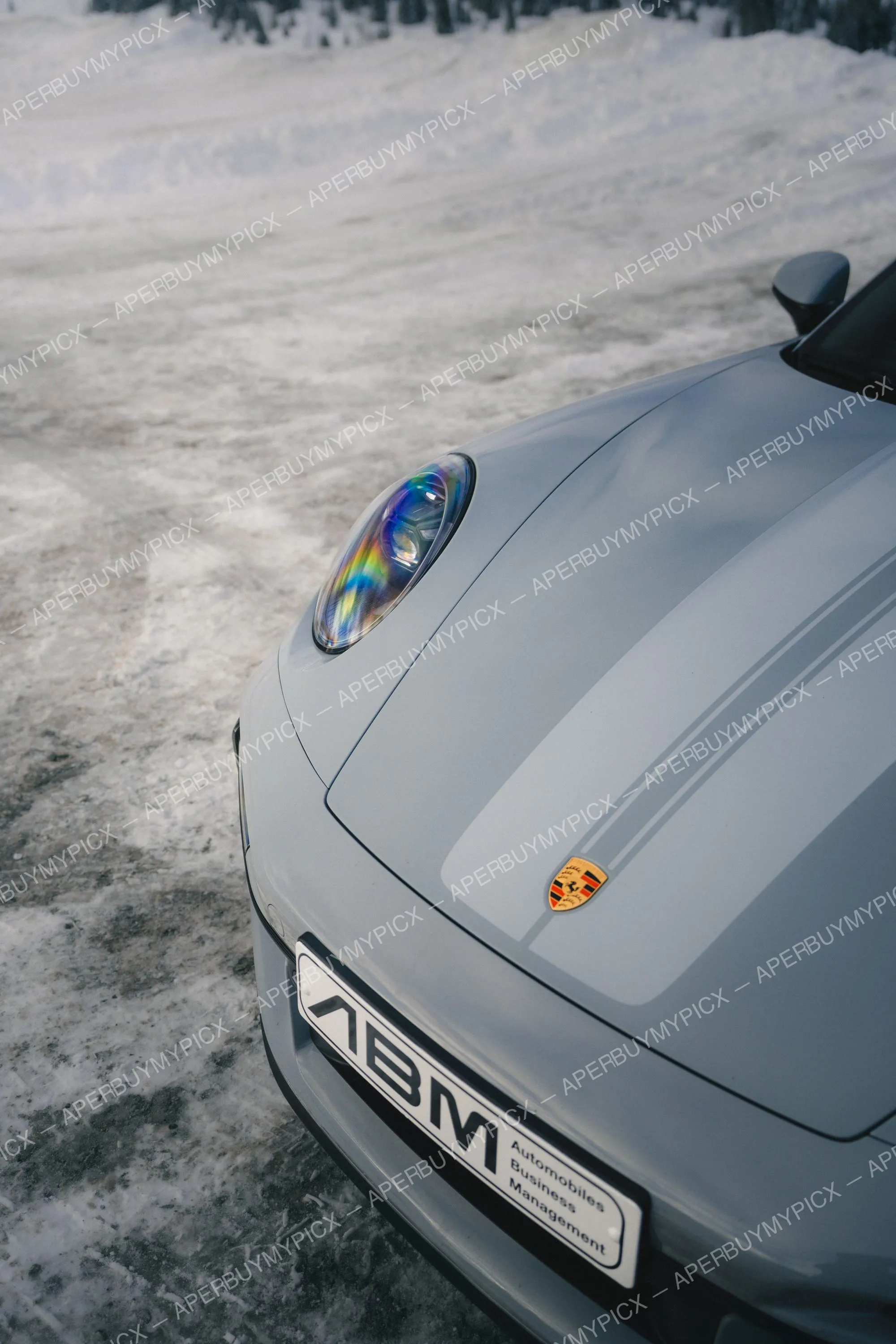 Avant d'une Porsche 911 grise avec écusson et phare aux reflets irisés, garée sur un sol enneigé à Megève, plaque ABM visible.