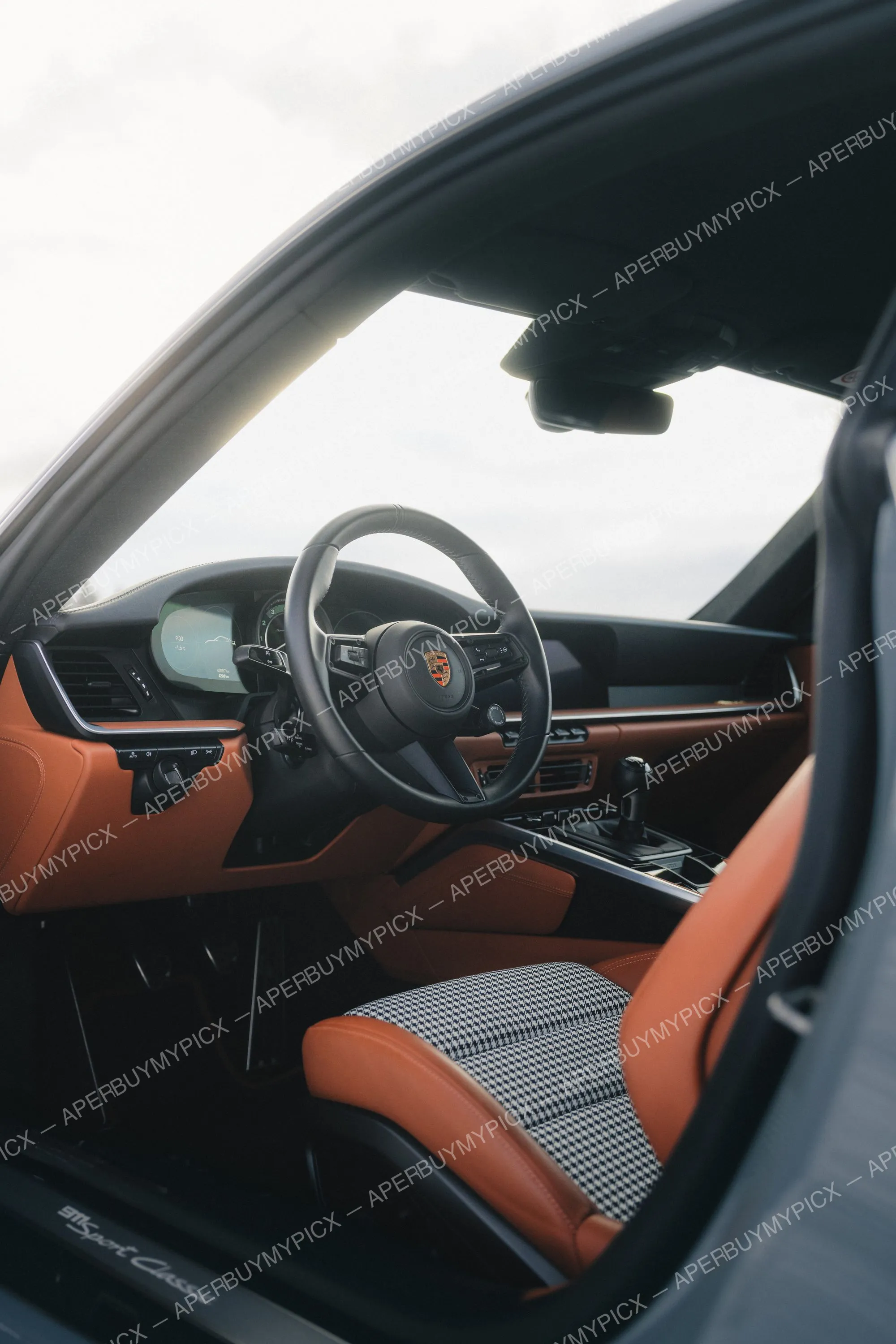 Intérieur d'une Porsche 911 Sport Classic avec volant noir, sièges en cuir cognac et tissu pied-de-poule, levier de vitesses manuel et tableau de bord numérique.