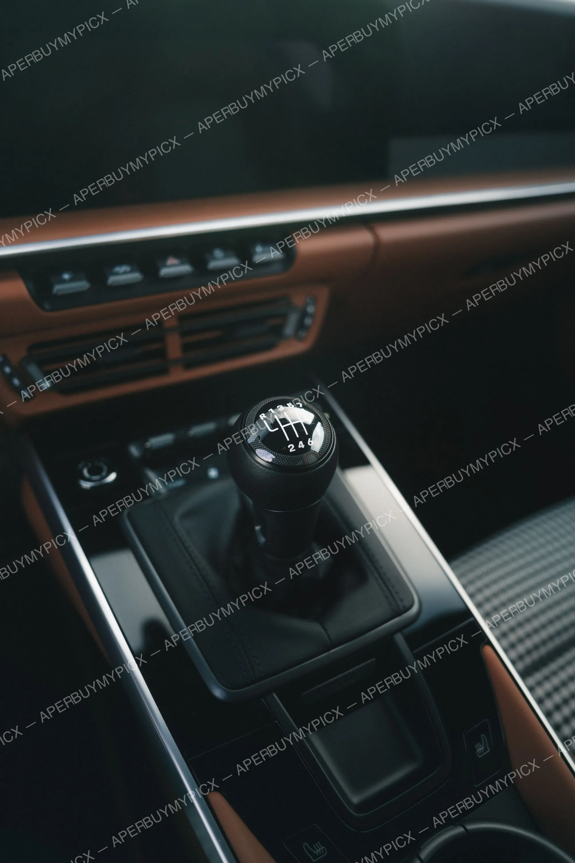 Gros plan sur un pommeau de levier de vitesses manuel à 7 rapports dans une voiture, avec console centrale noire, cuir cognac, aérateurs et boutons de commande en arrière-plan.