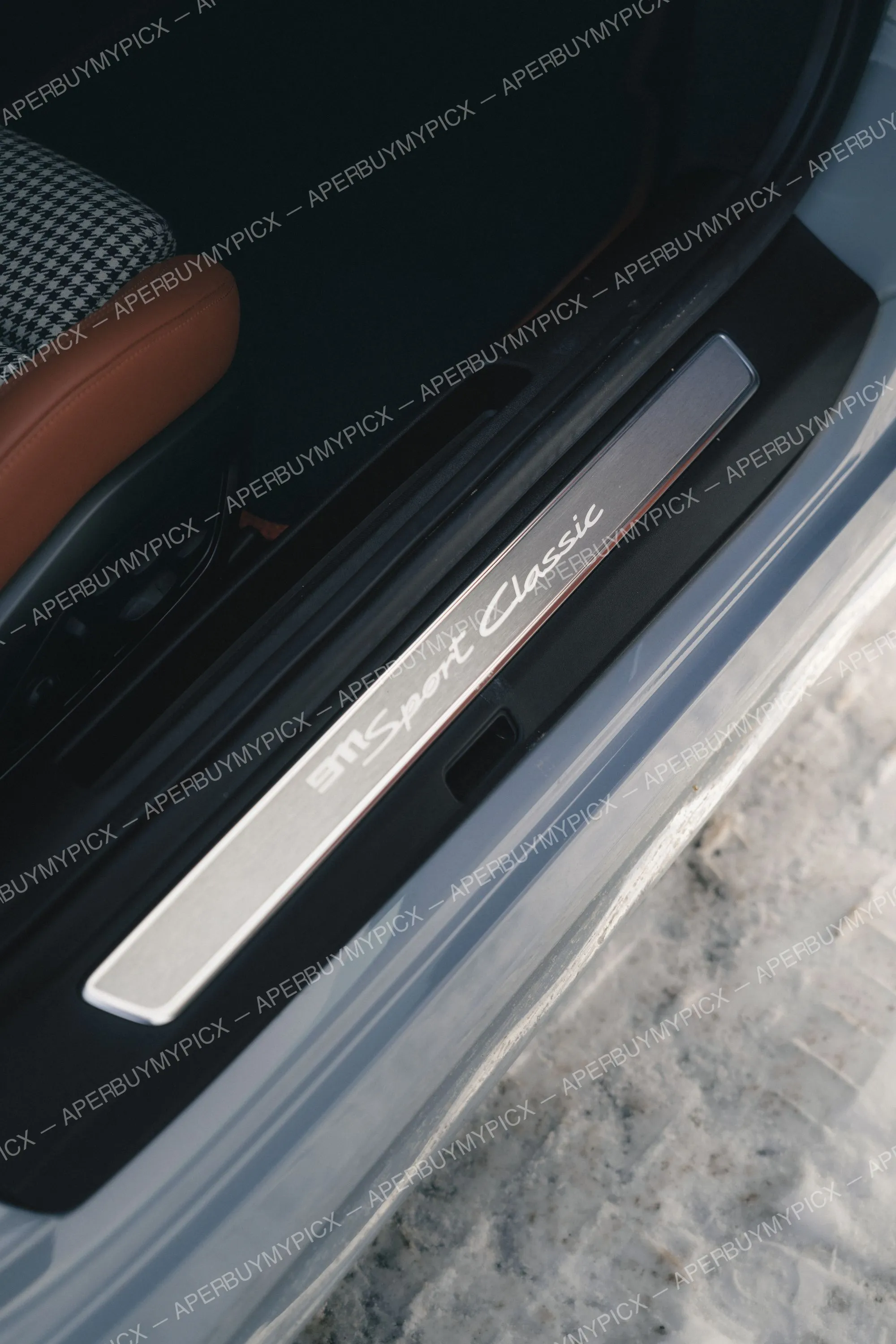 Gros plan sur le seuil de porte en aluminium brossé gravé « 911 Sport Classic » d'une Porsche, avec sièges en cuir brun cognac et tissu pied-de-poule visibles.