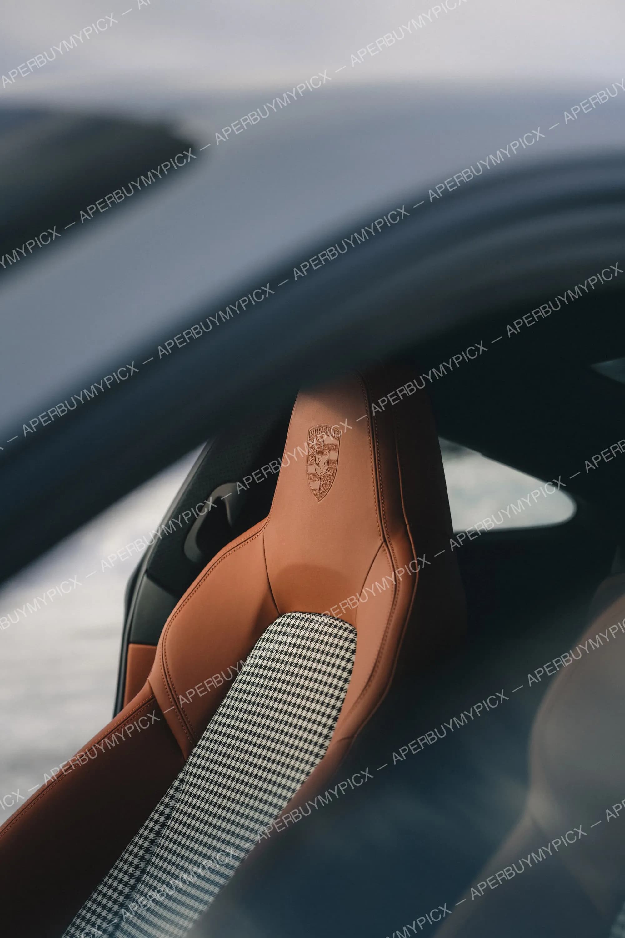 Gros plan sur un siège baquet Porsche en cuir cognac avec écusson gravé et centre en tissu pied-de-poule, vu à travers la vitre latérale.