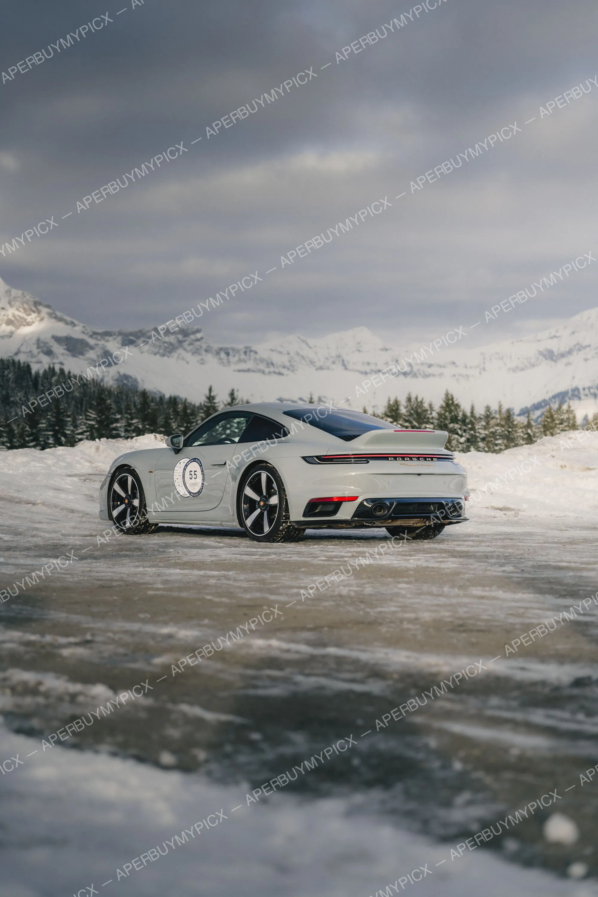 Porsche 911 Sport Classic grise portant le numéro 55 stationnée sur une piste enneigée avec montagnes des Alpes et forêt de sapins en arrière-plan, vue arrière trois-quarts.