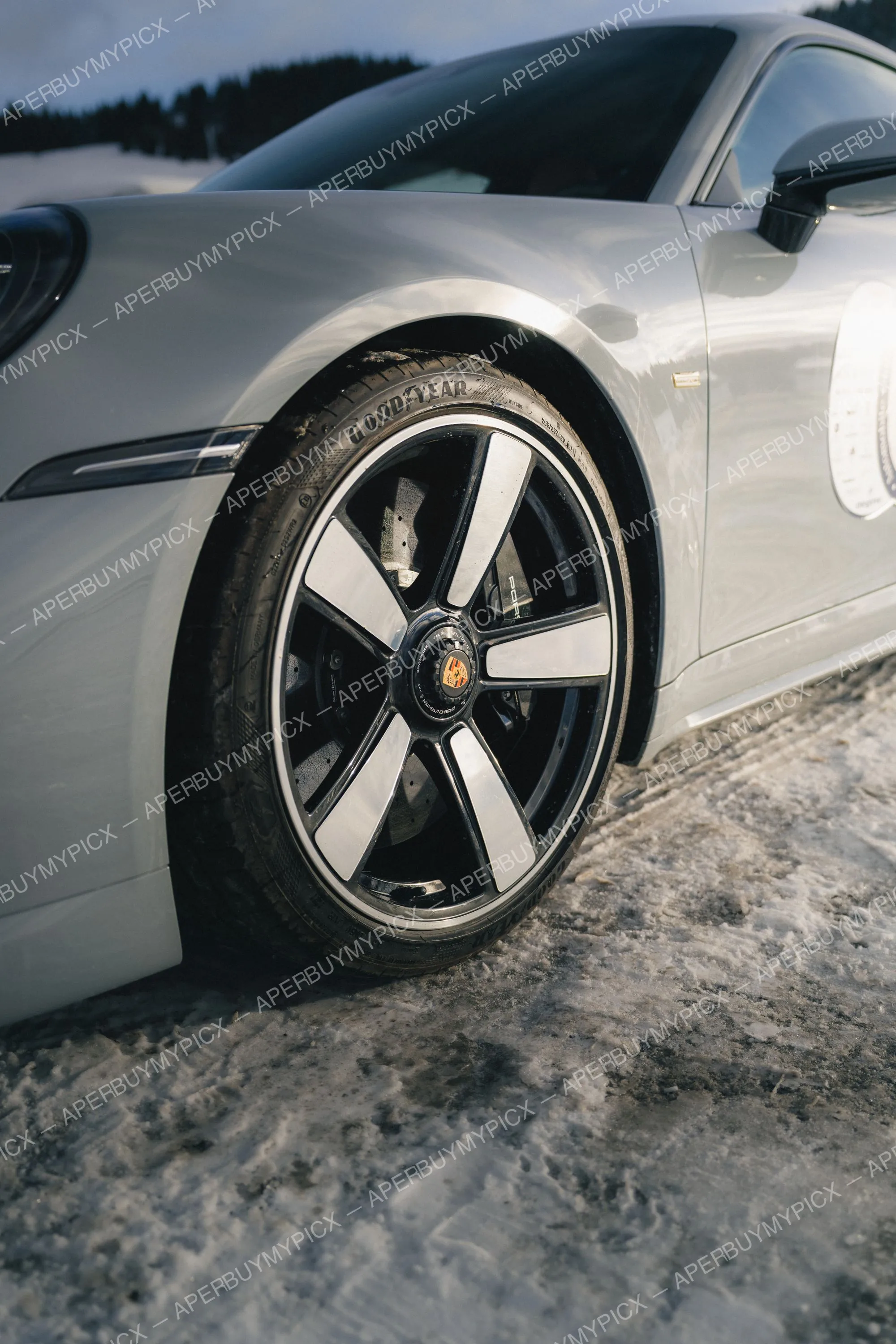 Gros plan sur la roue avant et l'aile d'une Porsche 911 grise avec jante noire et argent et pneu Goodyear, garée sur une route enneigée à Megève