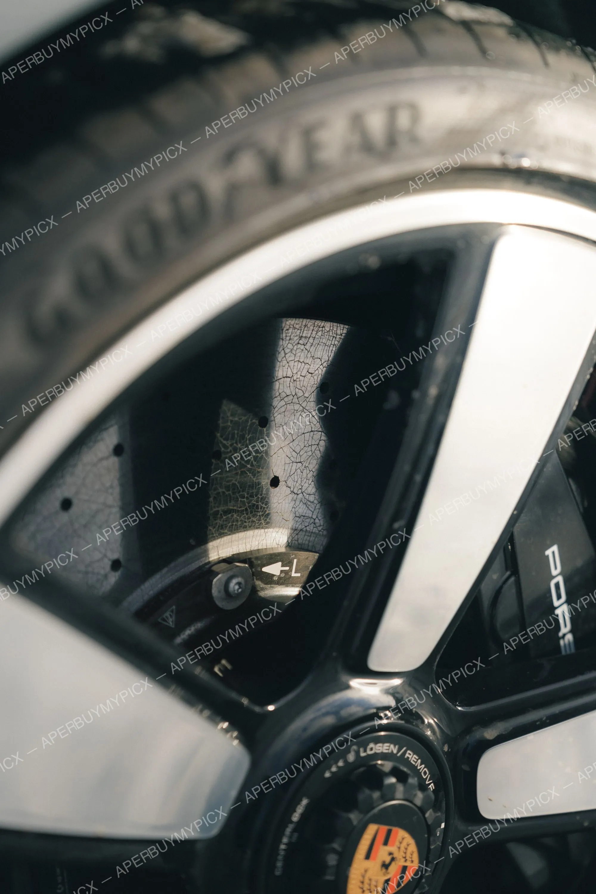 Gros plan sur une jante Porsche noire et argent avec pneu Goodyear, disque de frein céramique et écrou central marqué Lösen/Remove, logo Porsche visible.