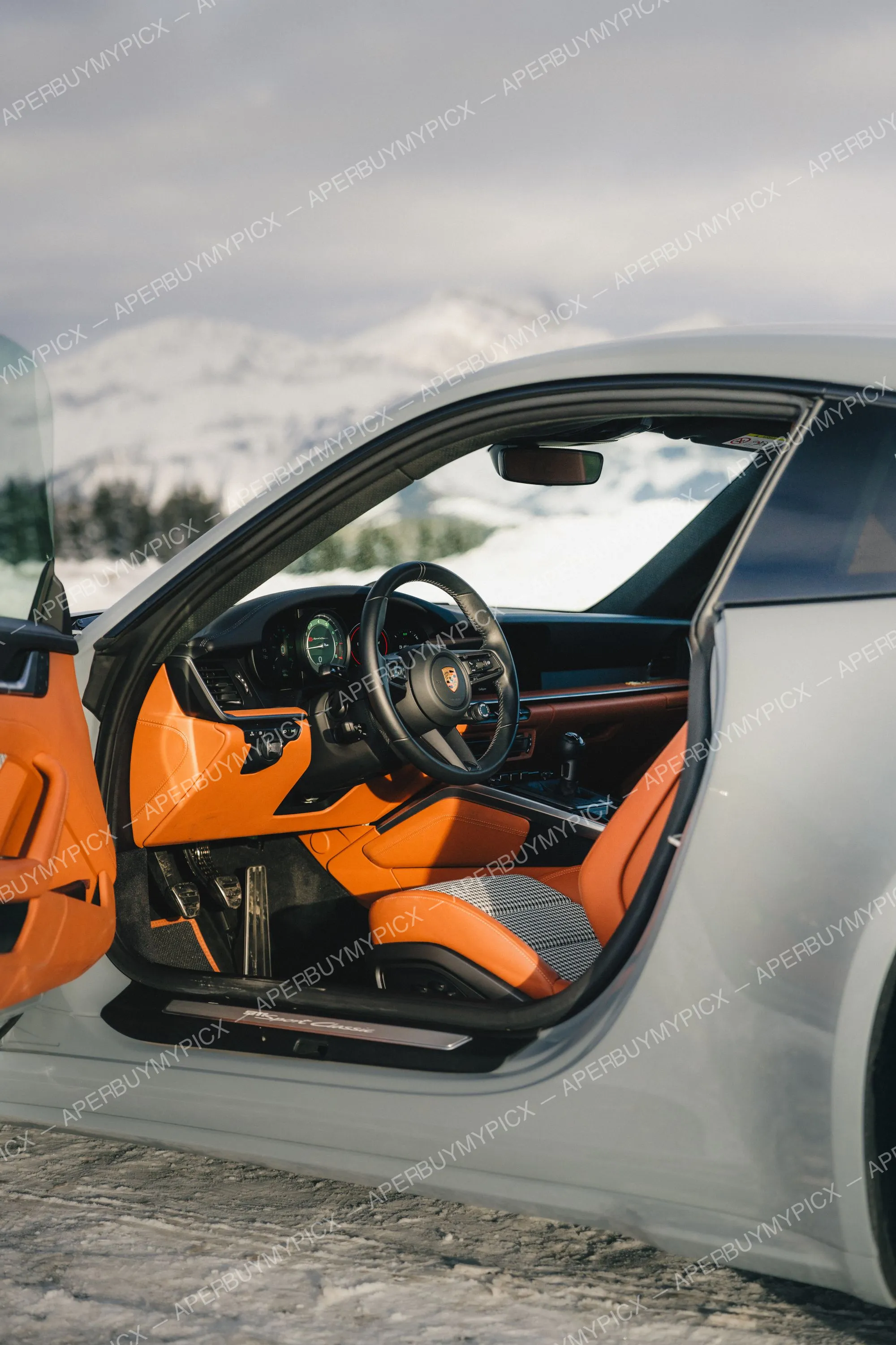 Intérieur d'une Porsche 911 Sport Classic argentée avec sièges en cuir orange, porte ouverte, devant un paysage de montagnes enneigées à Megève