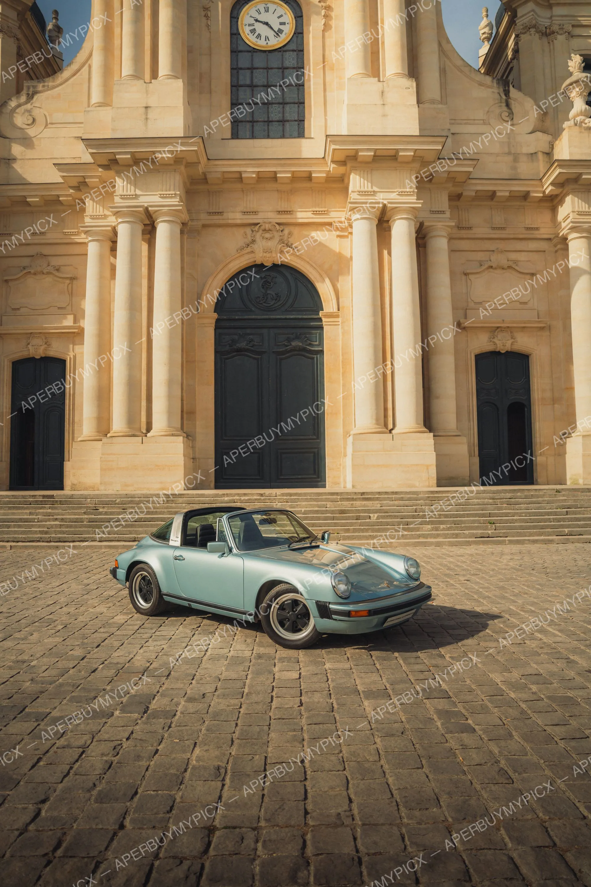 Porsche 911 SC Targa bleu clair garée sur des pavés devant la façade baroque d'une église avec horloge et grandes portes noires.