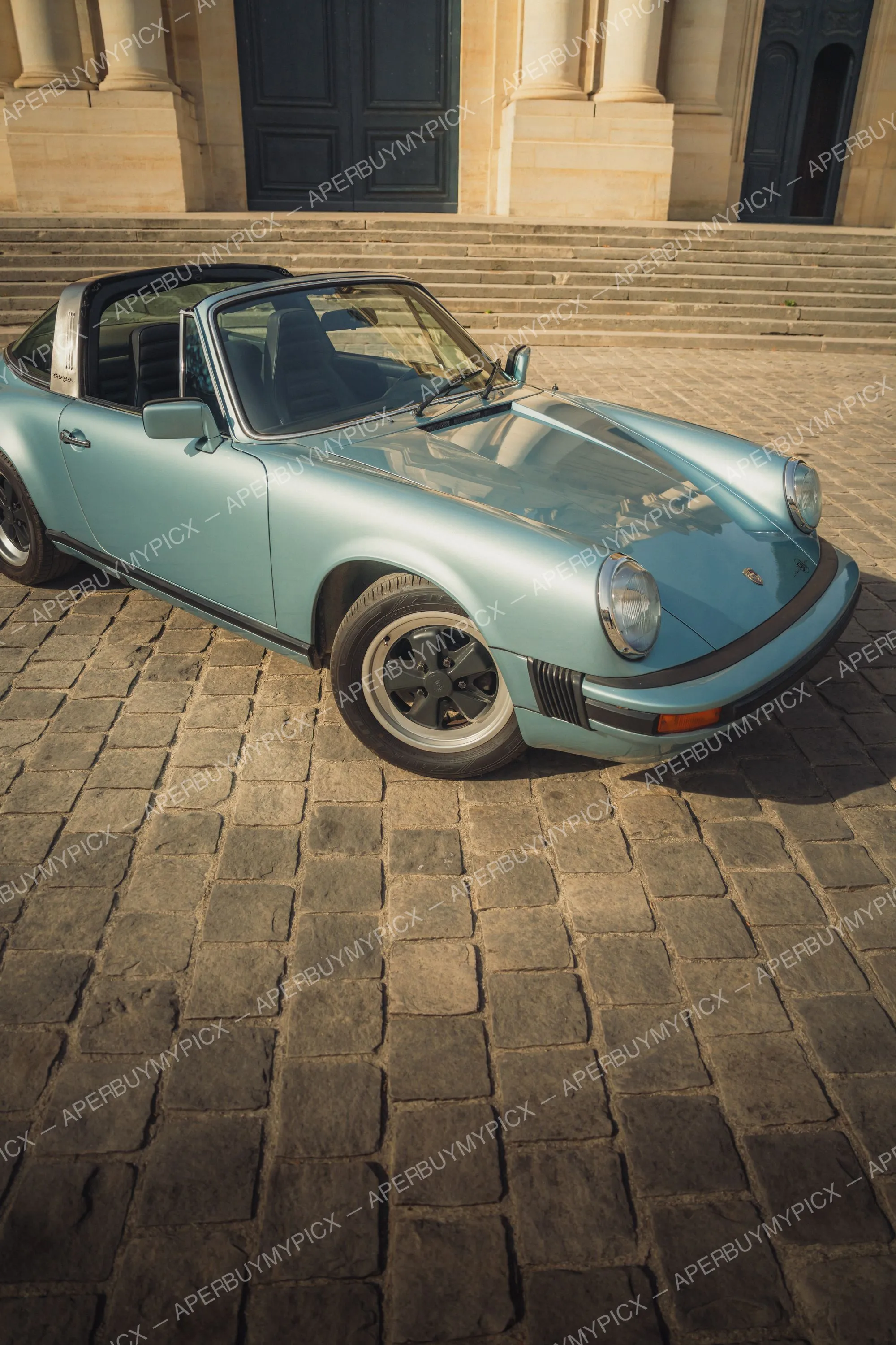 Porsche 911 SC Targa bleu clair garée sur une place pavée devant un bâtiment classique en pierre à Paris, capote ouverte, jantes Fuchs, au soleil couchant