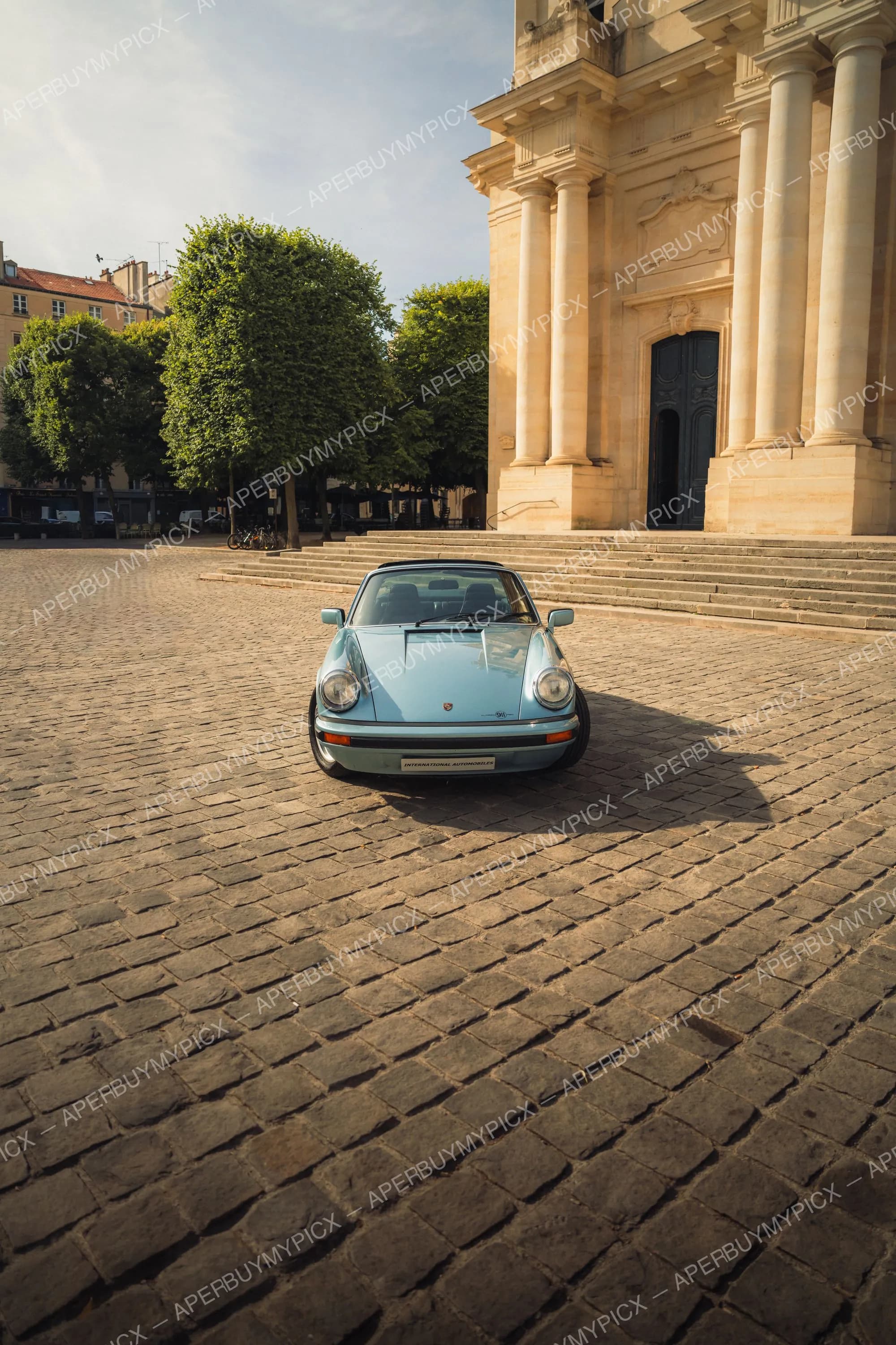 Porsche 911 SC Targa bleu clair vue de face, garée sur une place pavée devant un édifice néoclassique à colonnes au coucher du soleil.